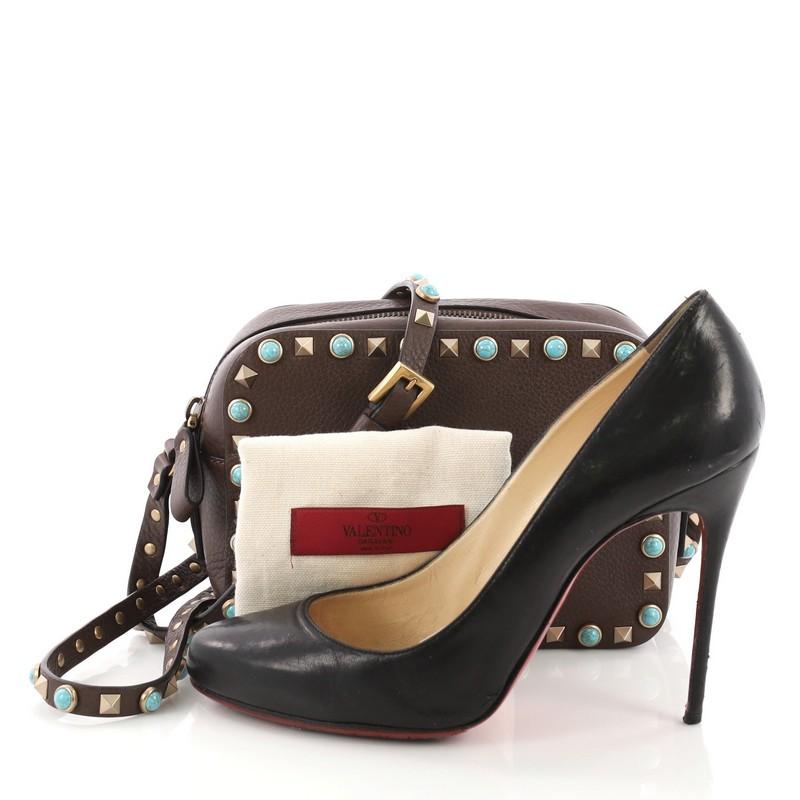 This Valentino Rockstud Camera Crossbody Bag Leather with Cabochons, crafted from brown leather with cabochons, features an adjustable crossbody strap, signature pyramid rockstud detailing, and gold-tone hardware. Its zip closure opens to a brown