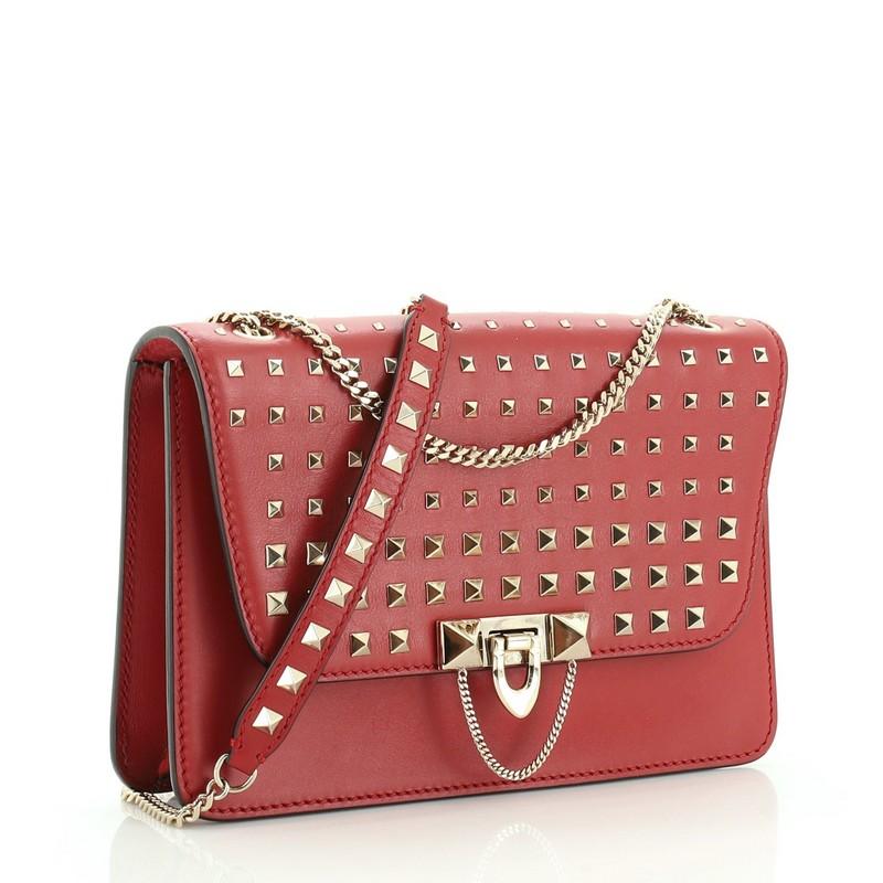 This Valentino Rockstud Demilune Shoulder Bag Studded Leather Small, crafted from red studded leather, features chain link strap with studded leather pad, exterior back slip pocket, and gold-tone hardware. Its flip-clasp closure opens to a red suede