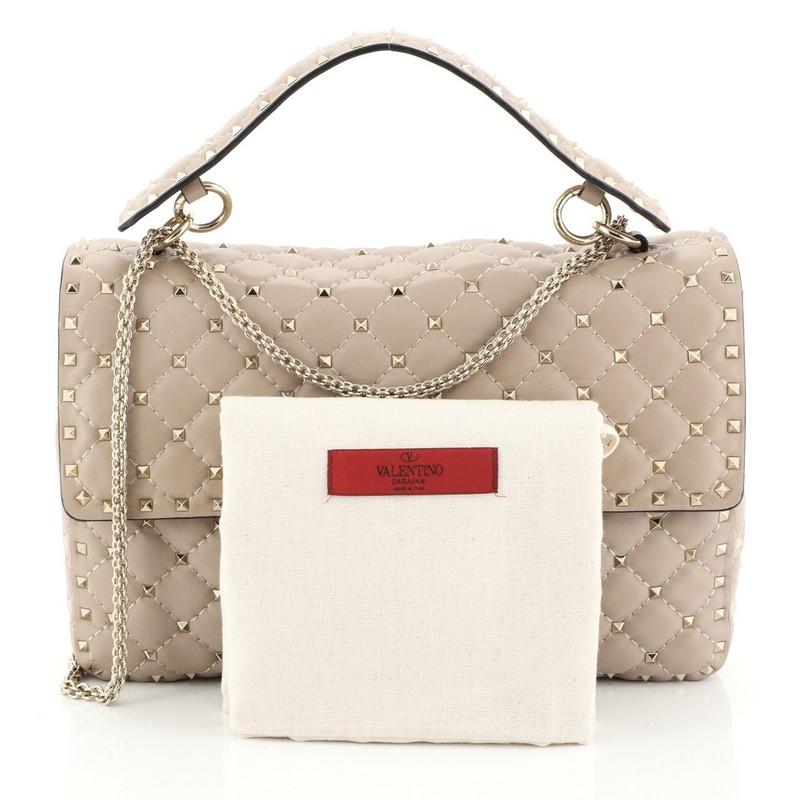 This Valentino Rockstud Spike Flap Bag Quilted Leather Large, crafted from neutral quilted leather, features a leather top handle, chain link strap, pyramid stud detailing, and gold-tone hardware. Its turn-lock closure opens to a neutral leather