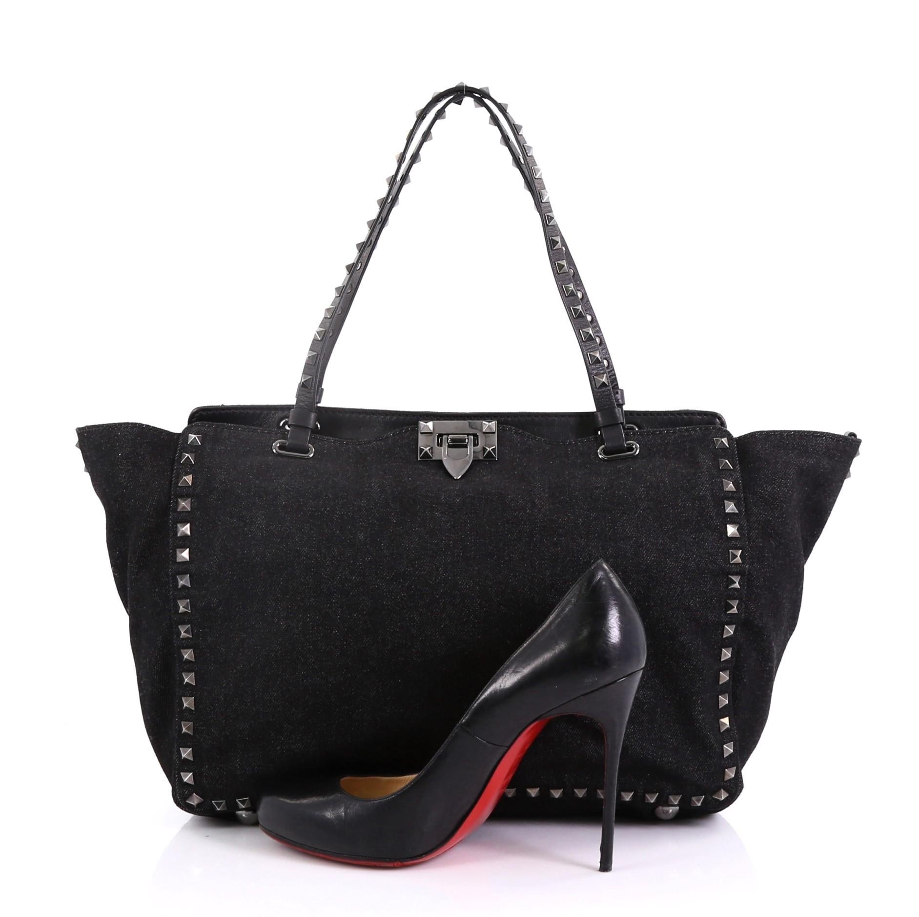 This Valentino Rockstud Tote Denim Medium, crafted in black denim, features dual studded handles, pyramid stud borders, and gunmetal-tone hardware. Its flip-clasp and top zip closures open to a black fabric interior with zip and slip pockets.