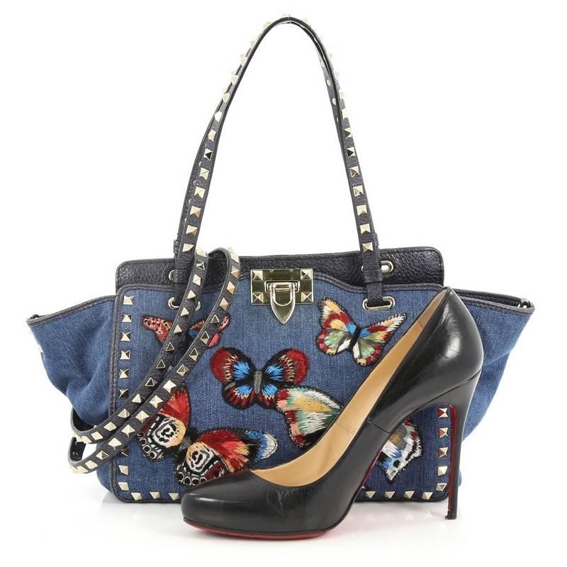 This authentic Valentino Rockstud Tote Denim with Butterfly Applique Small mixes edgy style with luxurious detailing. Crafted from blue denim with butterfly applique, this stylish tote features dual tall flat handles, gold-tone pyramid stud trim