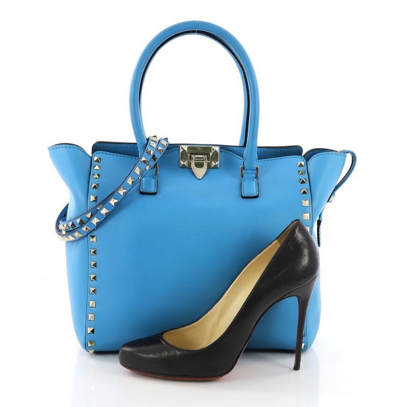 This Valentino Rockstud Tote Rigid Leather Medium, crafted in blue rigid leather, features dual rolled handles, pyramid stud borders, and gold-tone hardware. Its flip-clasp and top zip closures open to a beige fabric interior with zip and slip