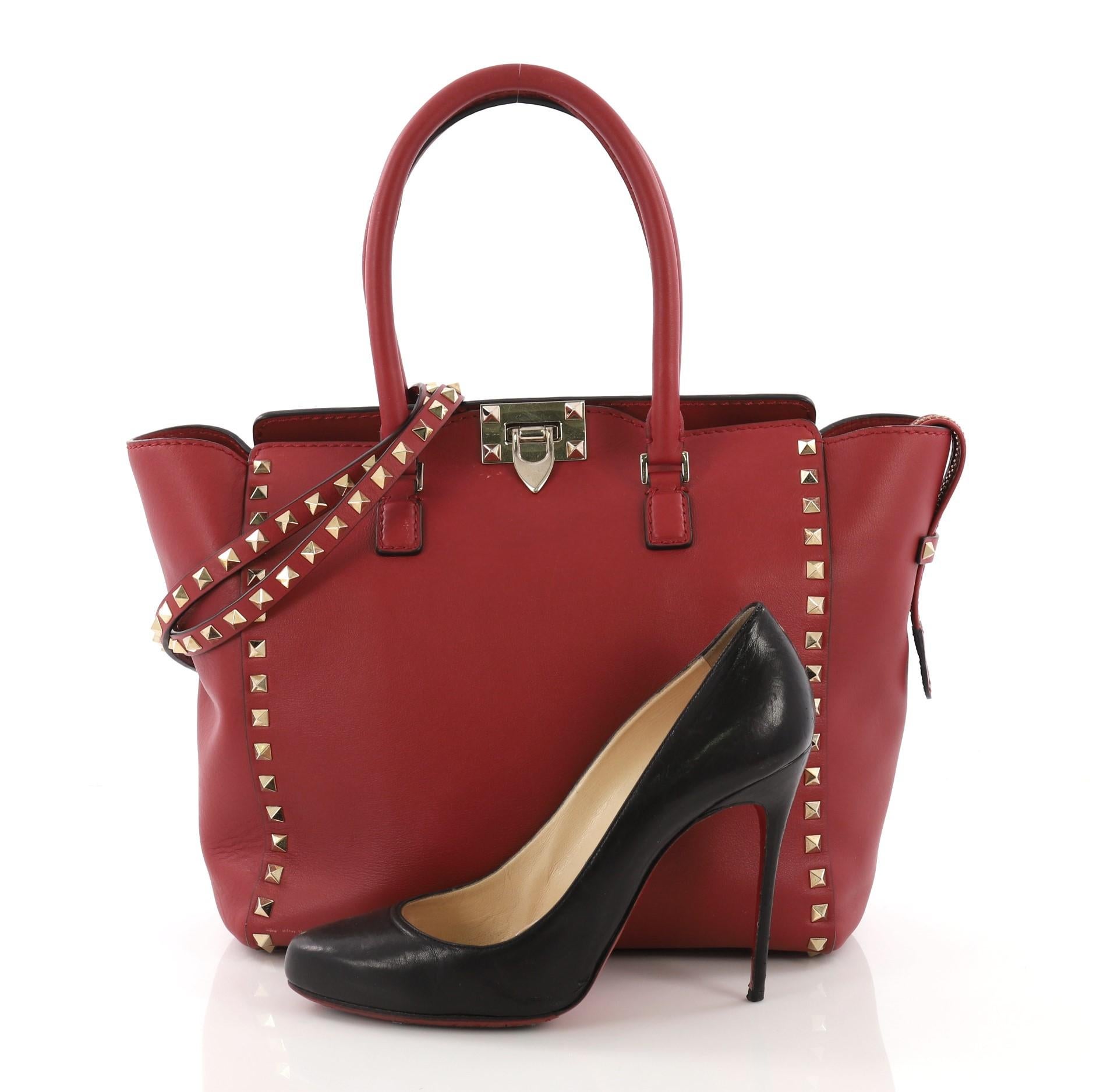 This Valentino Rockstud Tote Rigid Leather Medium, crafted in red rigid leather, features dual rolled handles, pyramid stud borders, and gold-tone hardware. Its flip-clasp and top zip closures open to a beige fabric interior with zip and slip