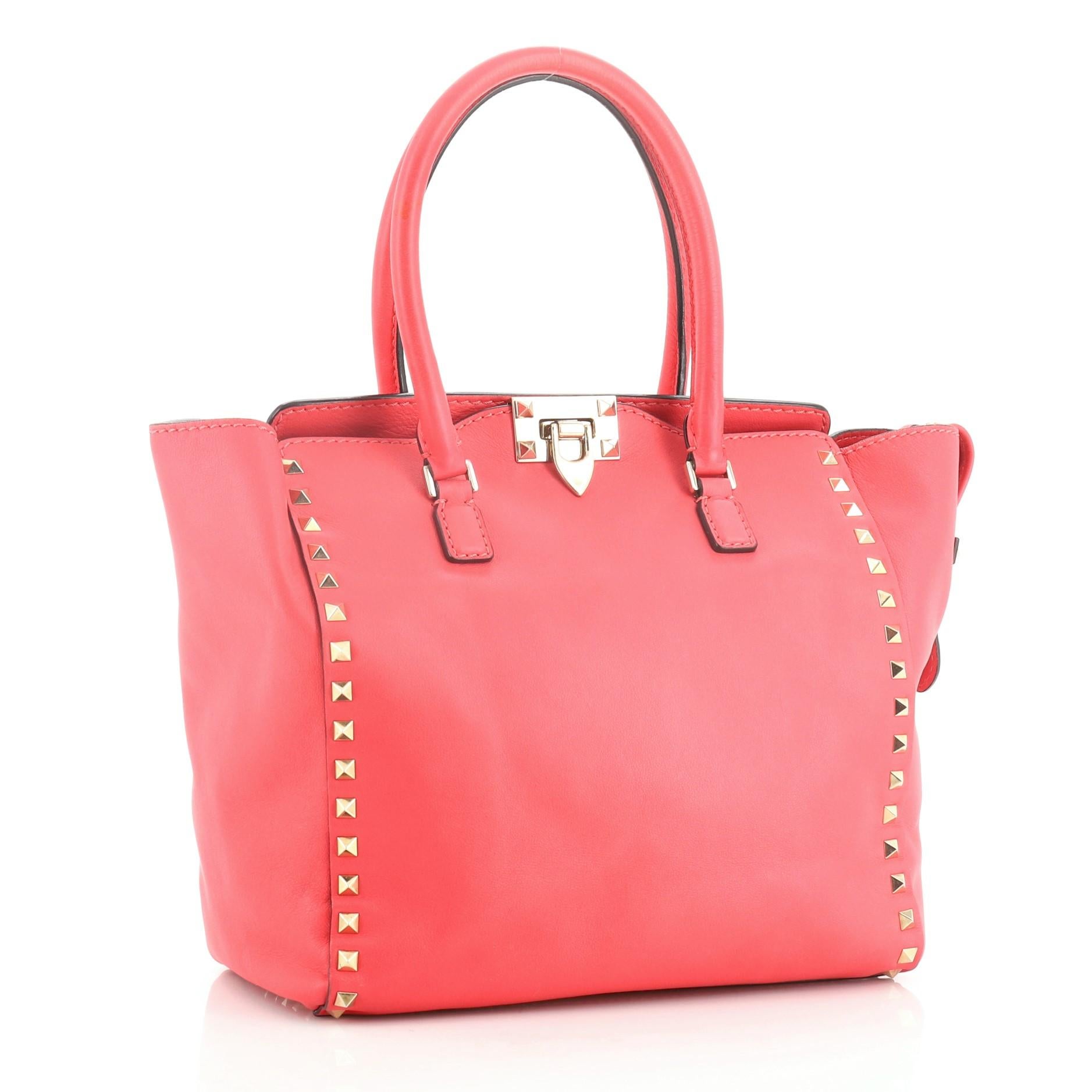 This Valentino Rockstud Tote Rigid Leather Medium, crafted from pink leather, features signature pyramid stud border, dual-rolled handles, and gold-tone hardware. Its flip-clasp and top zip closures open to a neutral fabric interior with side zip