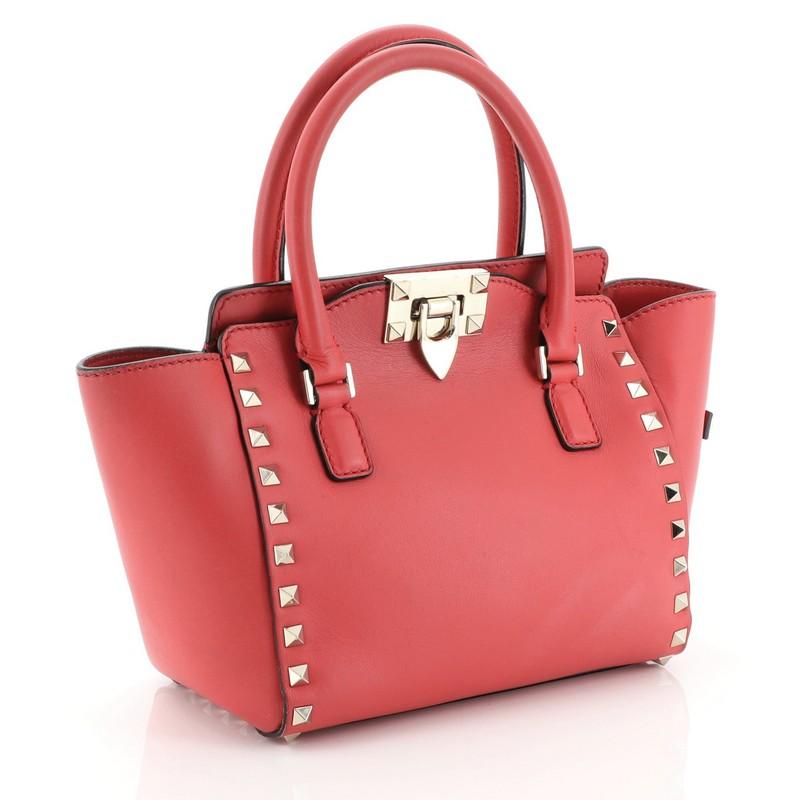 This Valentino Rockstud Tote Rigid Leather Mini, crafted from red rigid leather, features pyramid stud border, dual rolled handles, and gold-tone hardware. Its flip-clasp and zip closure opens to a neutral fabric interior with zip and slip pockets.