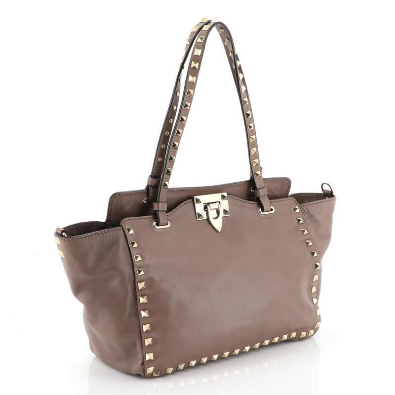 This Valentino Rockstud Tote Soft Leather Small, crafted from neutral leather, features dual tall flat handles, pyramid stud trim details, and gold-tone hardware. Its clasp lock closure opens to a neutral fabric interior with side zip pocket.