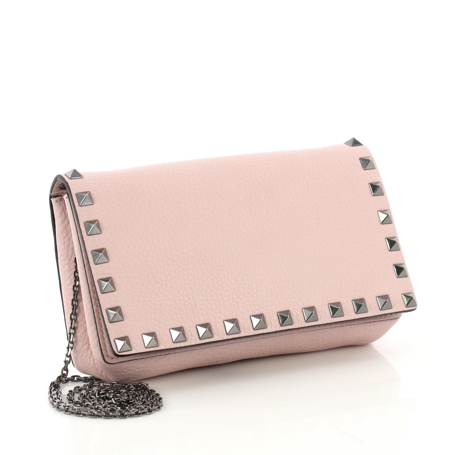 This Valentino Rockstud Wallet On Chain Leather, crafted from pink leather, features pyramid stud embellishments, chain straps, and gunmetal-tone hardware. Its magnetic snap closure opens to a pink suede interior with multiple card slots, slip