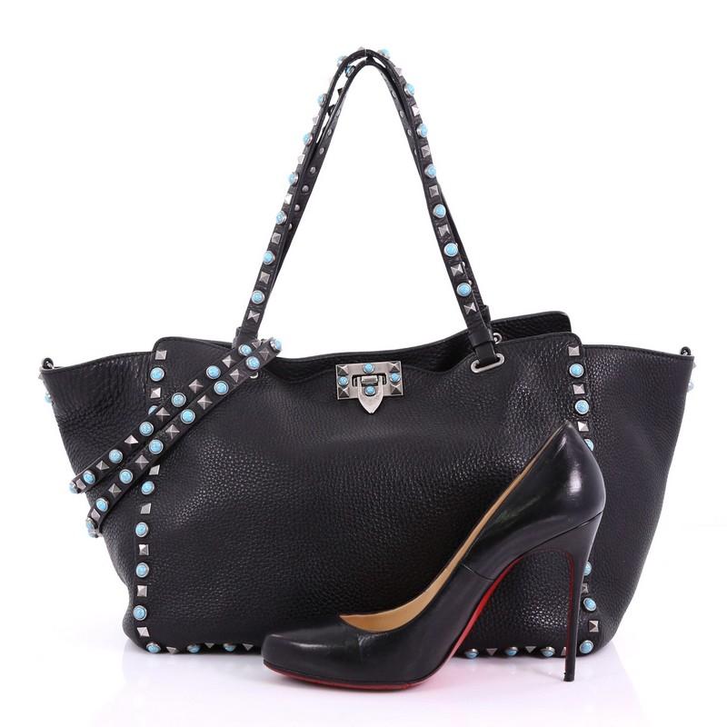 This Valentino Rolling Rockstud Tote Leather with Cabochons Medium, crafted from black leather, features studded dual-flat leather handles, signature rockstud and cabochon trims, and aged silver-tone hardware. Its flip-lock closure opens to a black