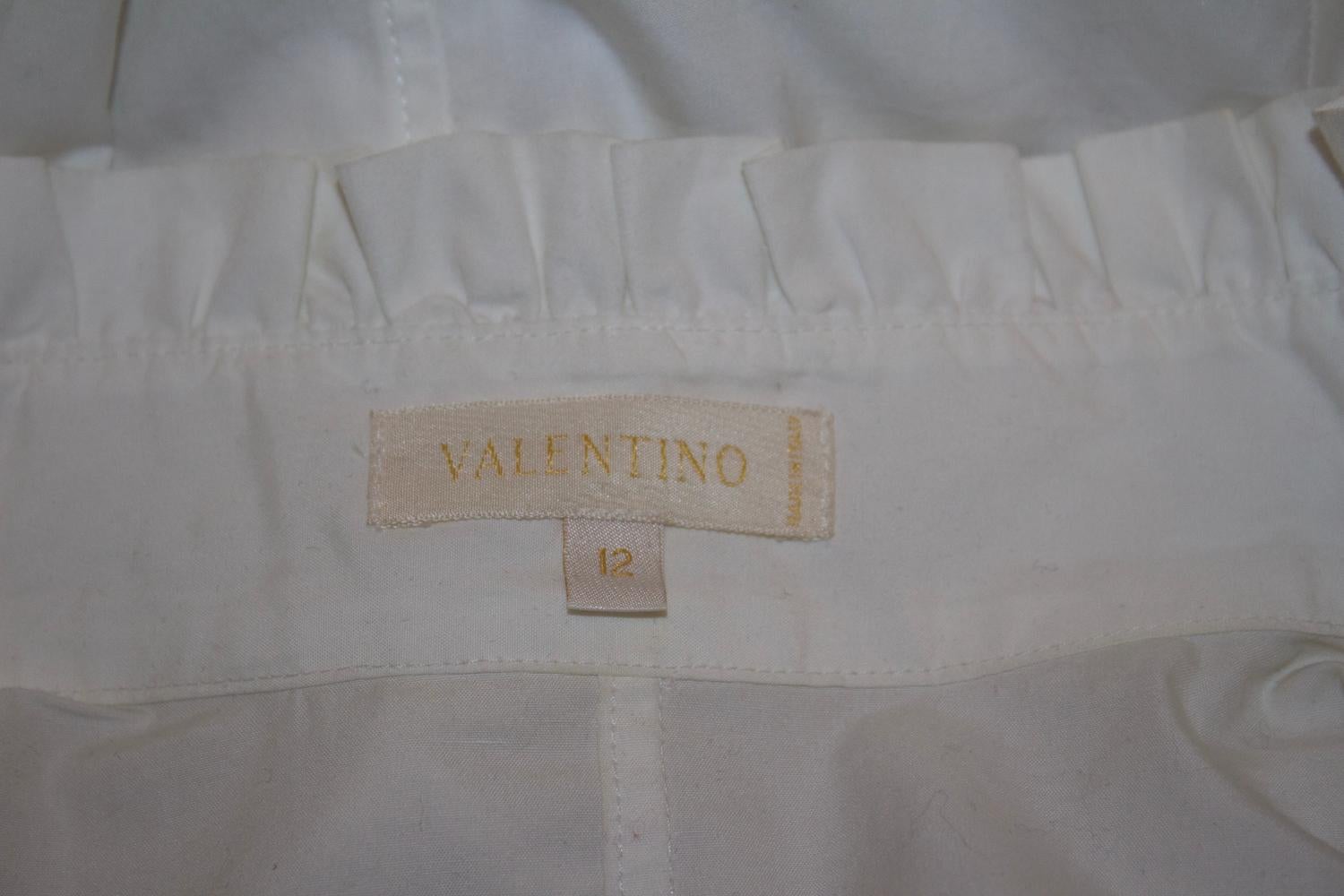 Una elegante camisa de algodón de Valentino, línea principal. En un magnífico algodón blanco, la camisa tiene el cuello alzado con un gran volante en la parte delantera. Tiene puños con botones y abertura delantera oculta con botones. 
Talla 12 del
