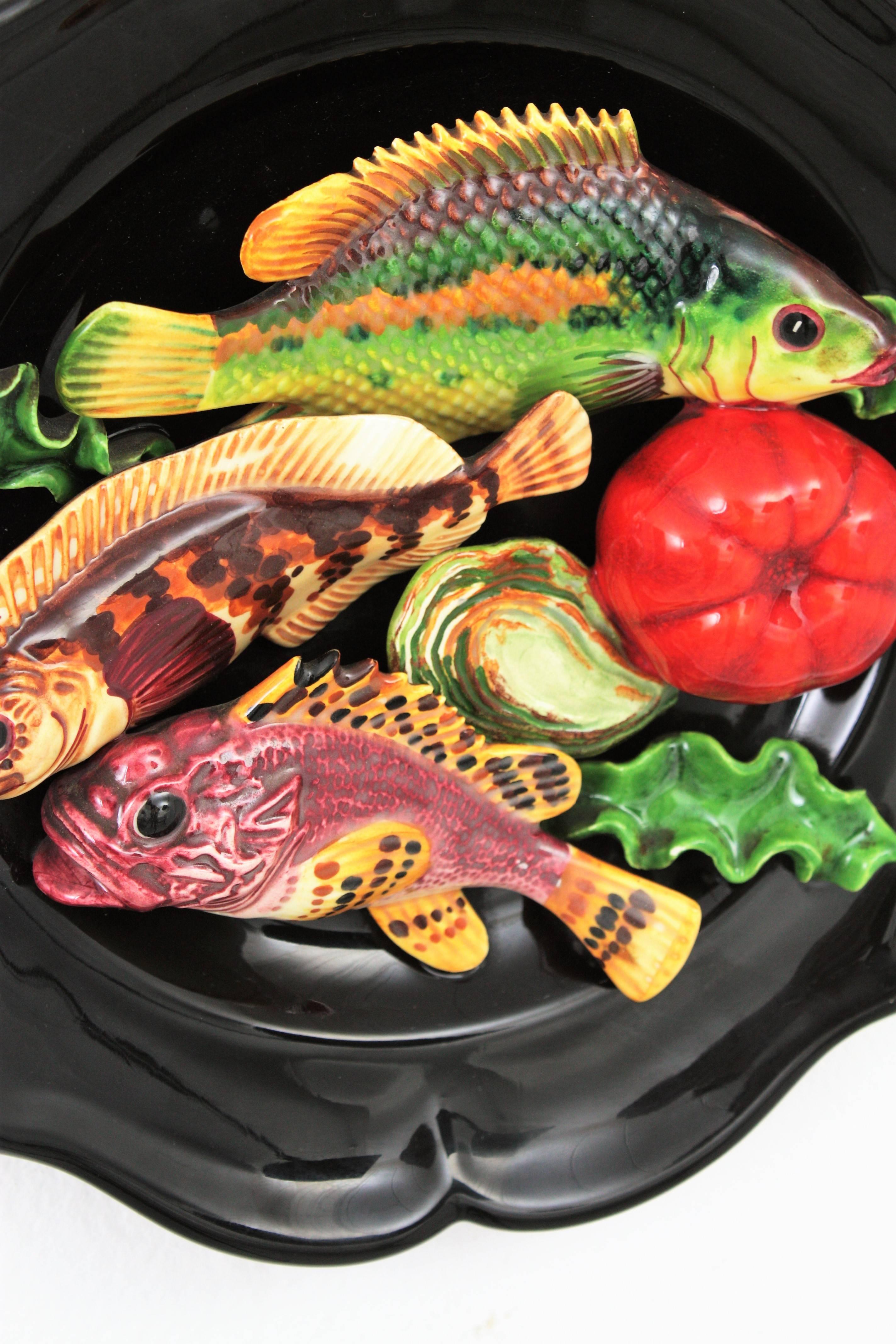 Attrayante assiette en trompe-l'œil en céramique émaillée Majolica attribuée à Vallauris. France, années 1960.
Cette grande assiette a un fond noir et un décor coloré en trompe-l'oeil composé de poissons, d'algues et de légumes. Cette plaque sera