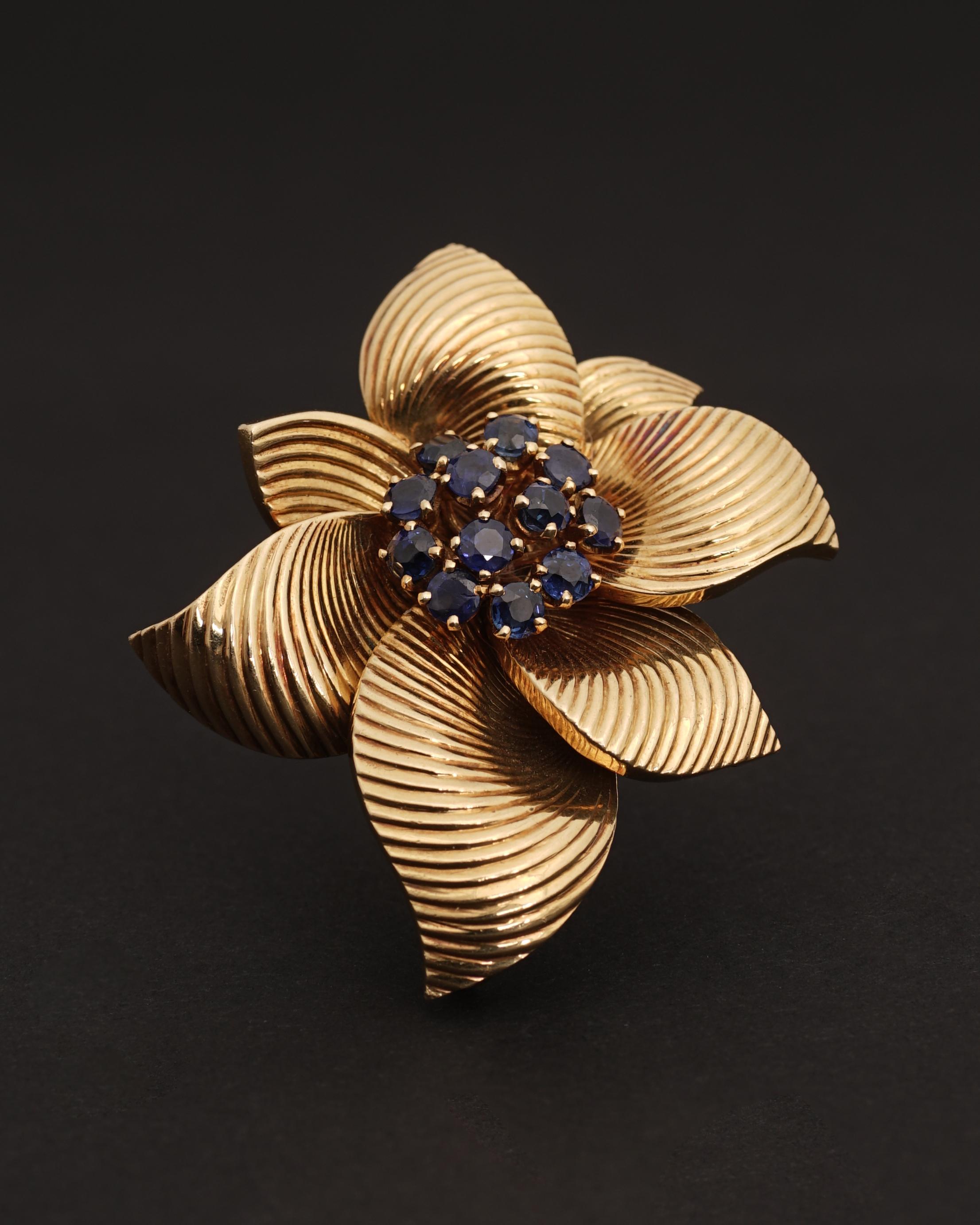 Van Cleef & Arpels, Manufactured by Pery & Fils.
Magnolia Brooch, Circa 1965.
18K Gold featuring 7 stylized petales and 12 round cushion blue sapphires.
Signed and Numbered Van Cleef & Arpels. Manufactured by Pery & Fils. 

The ruby version of this