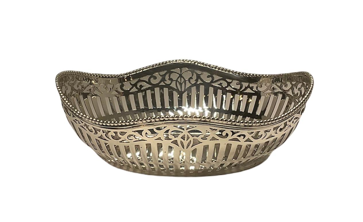 This an oval shaped bon bon silver basket. It has a serpentine shaped upper border. Its rim is decorated with a chain of beads. Its sides are adorned with reticulated vertical line and scrolls of foliage. Below the base is the Dutch hallmark of Van