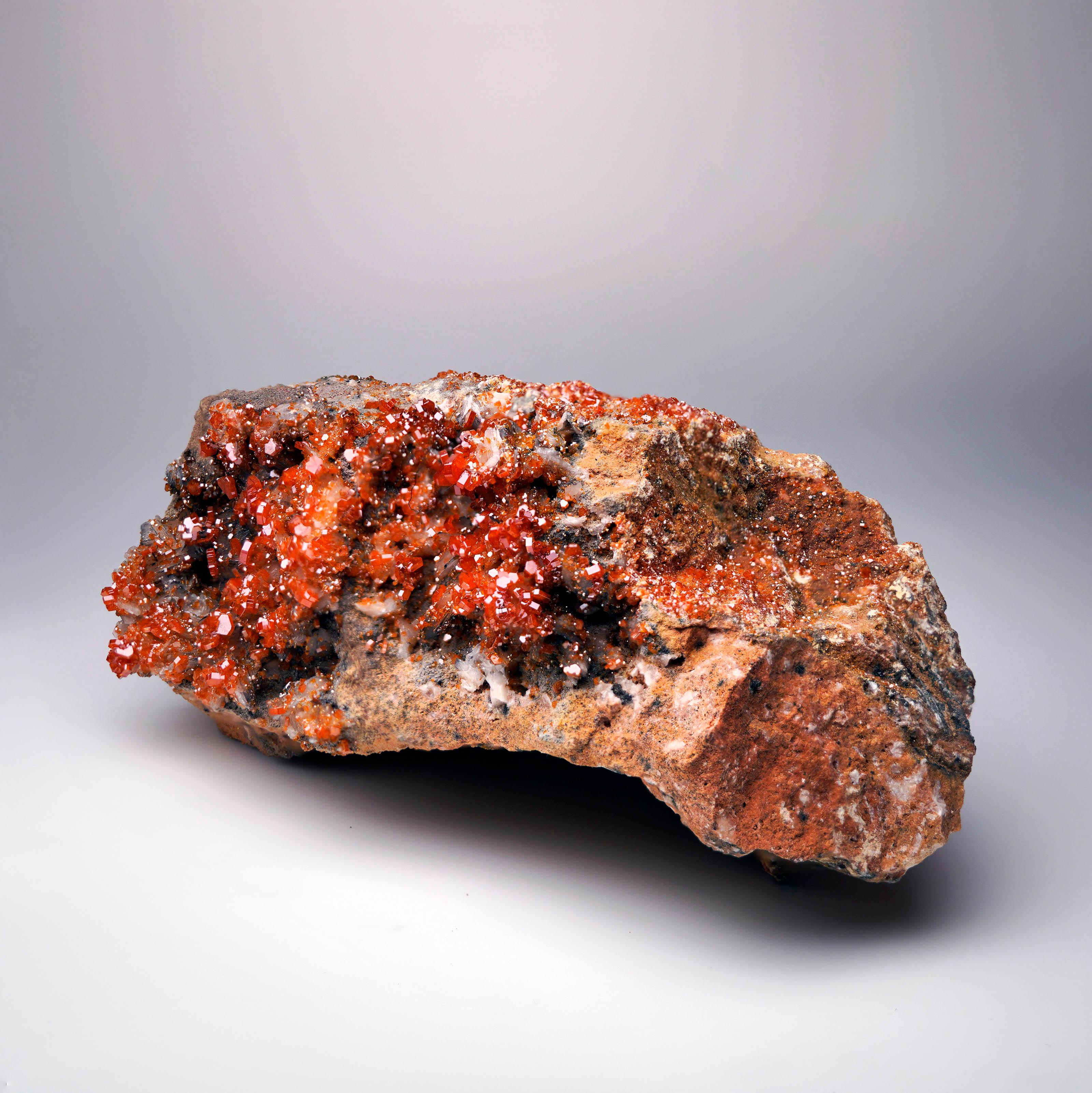 Este brillante espécimen muestra una reluciente superposición de cristales rojos de Vanadinita agrupados sobre una delicada y escultural Barita. El gran brillo y el color saturado de la Vanadinita llaman la atención frente a la Barita
