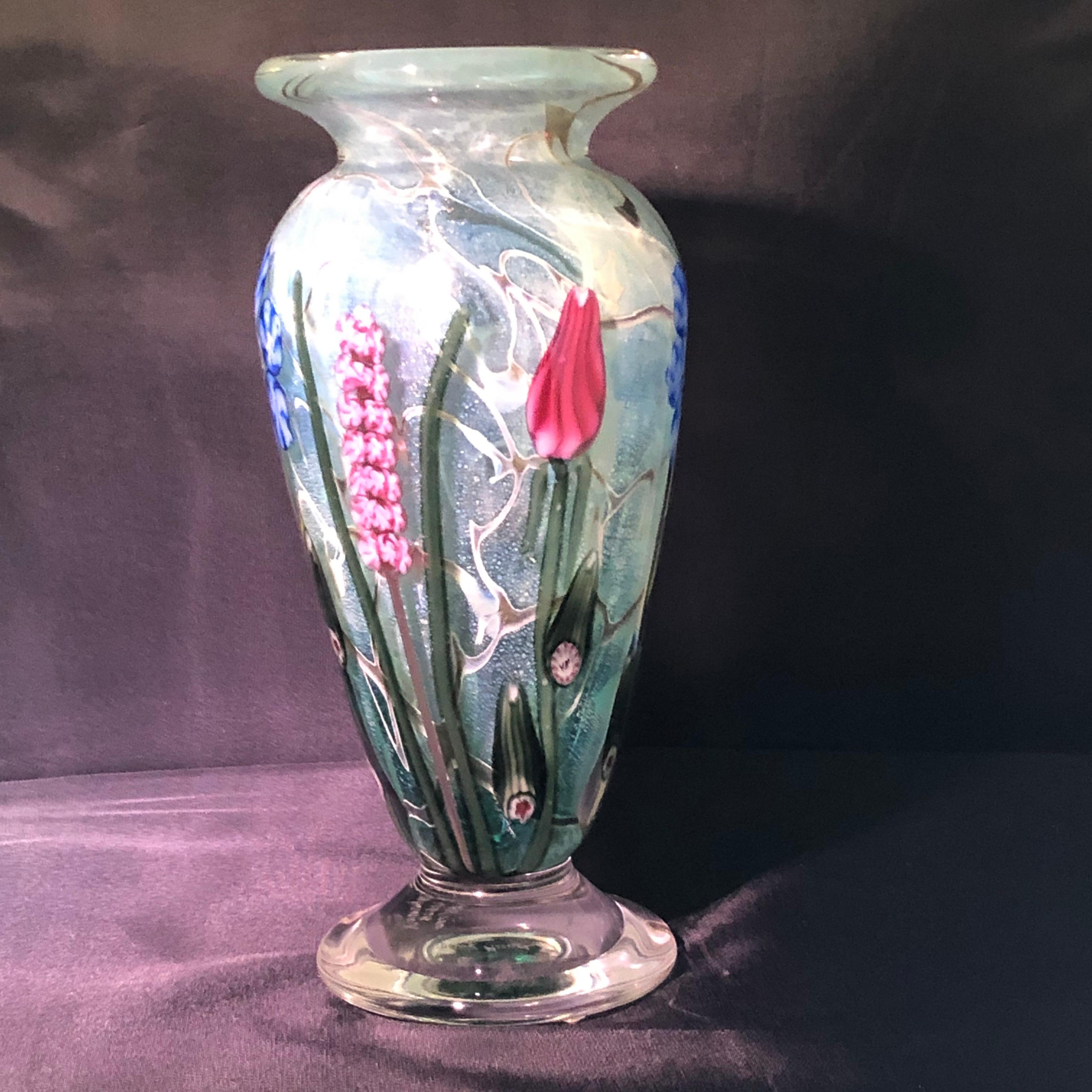 Hand blown Vandermark art glass vase decorated with outstanding bright colored spring flowers on a green colored background. It was hand blown so when the light hits the vase, you can see beautiful shimmering gold speckles throughout the vase. The