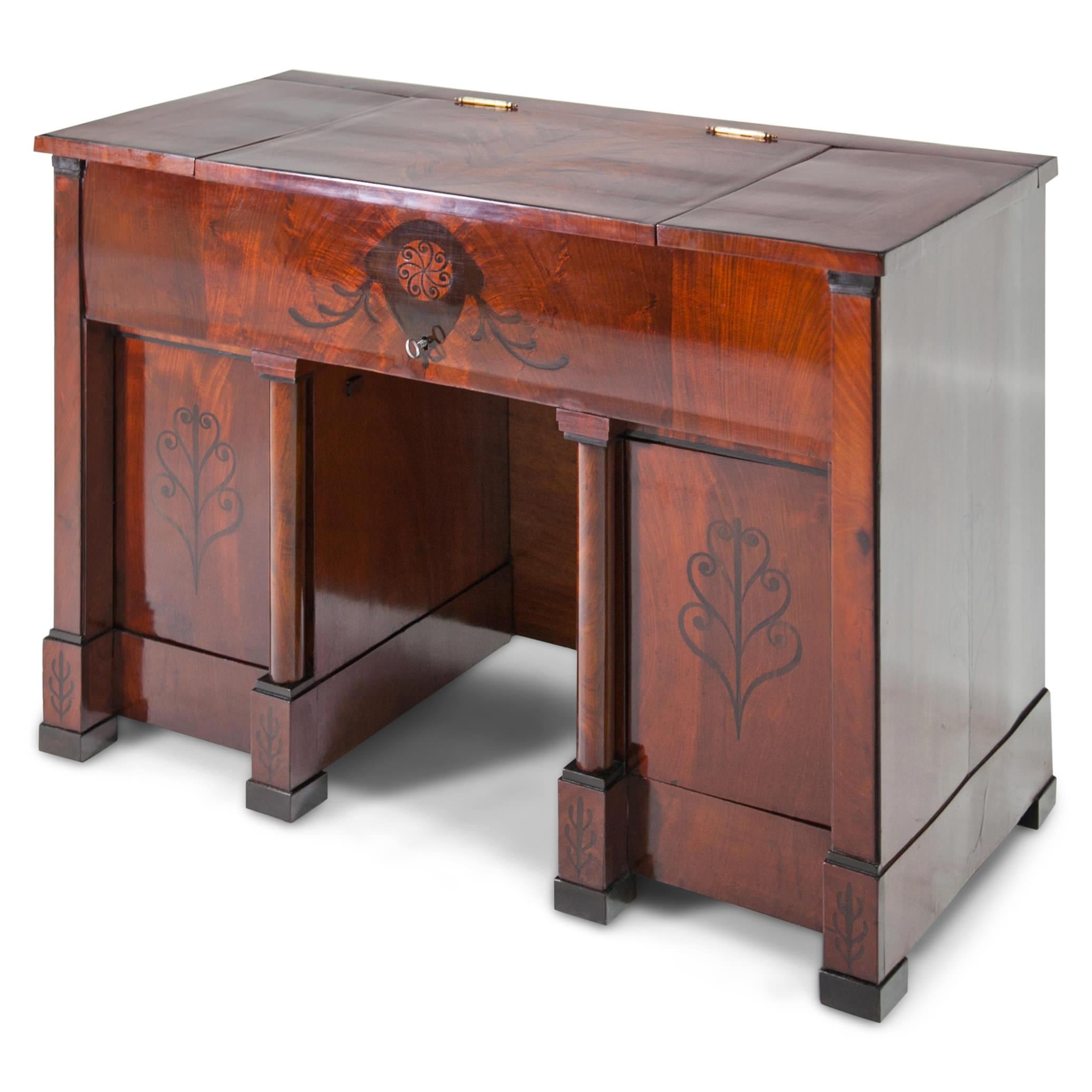 Mahogany veneered Biedermeier vanity desk with floral ornaments and pilaster as well as columns at the front with ebonized bases and capitals. The doors are opened via a pressure mechanism. The tabletop has a hinged lid with a mirror on the inside,