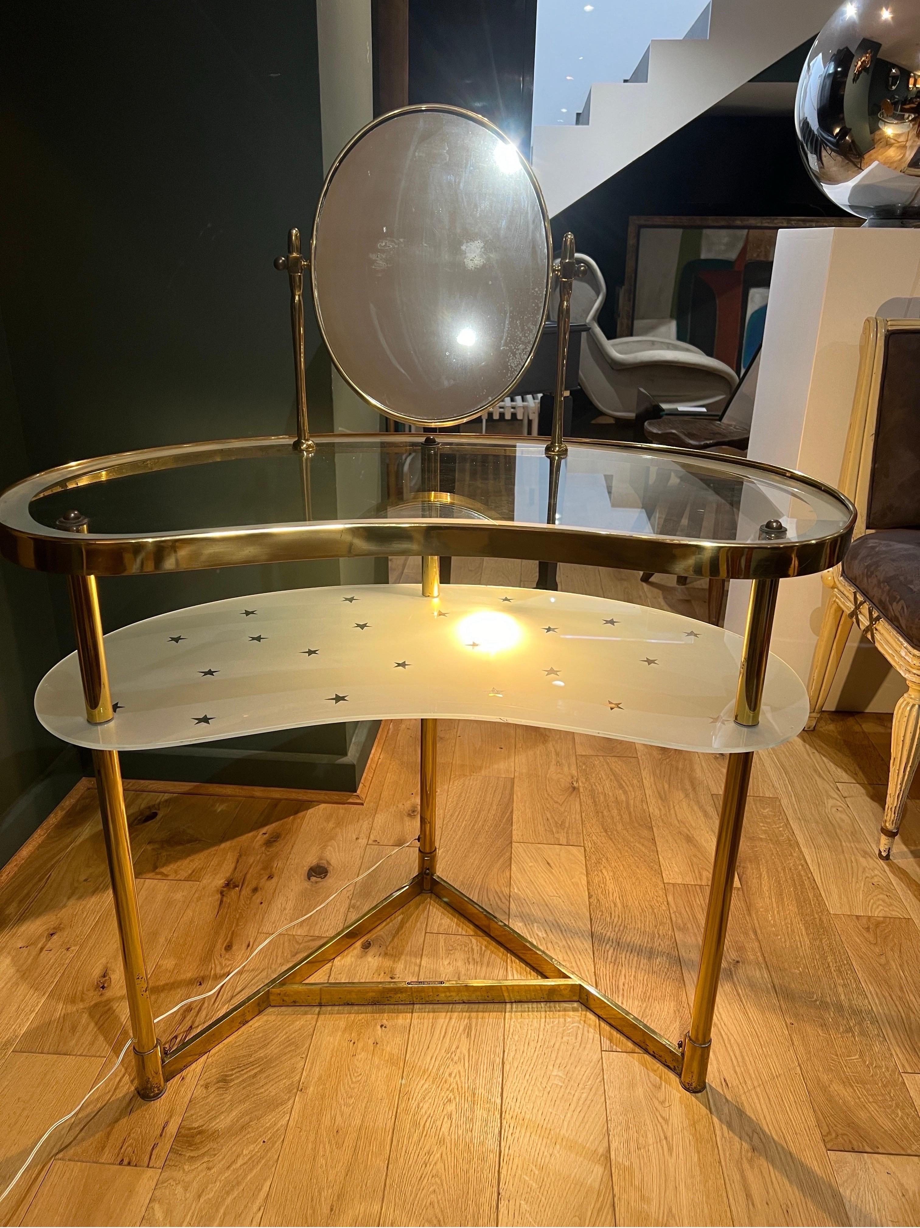 A Luigi Brusotti brass and glass kidney shaped vanity table . The table is composed of a swivel brass framed mirror mounted on a transparent glass top tier and frosted lower tier with etched stars decoration. 
There’s a light fixture under the