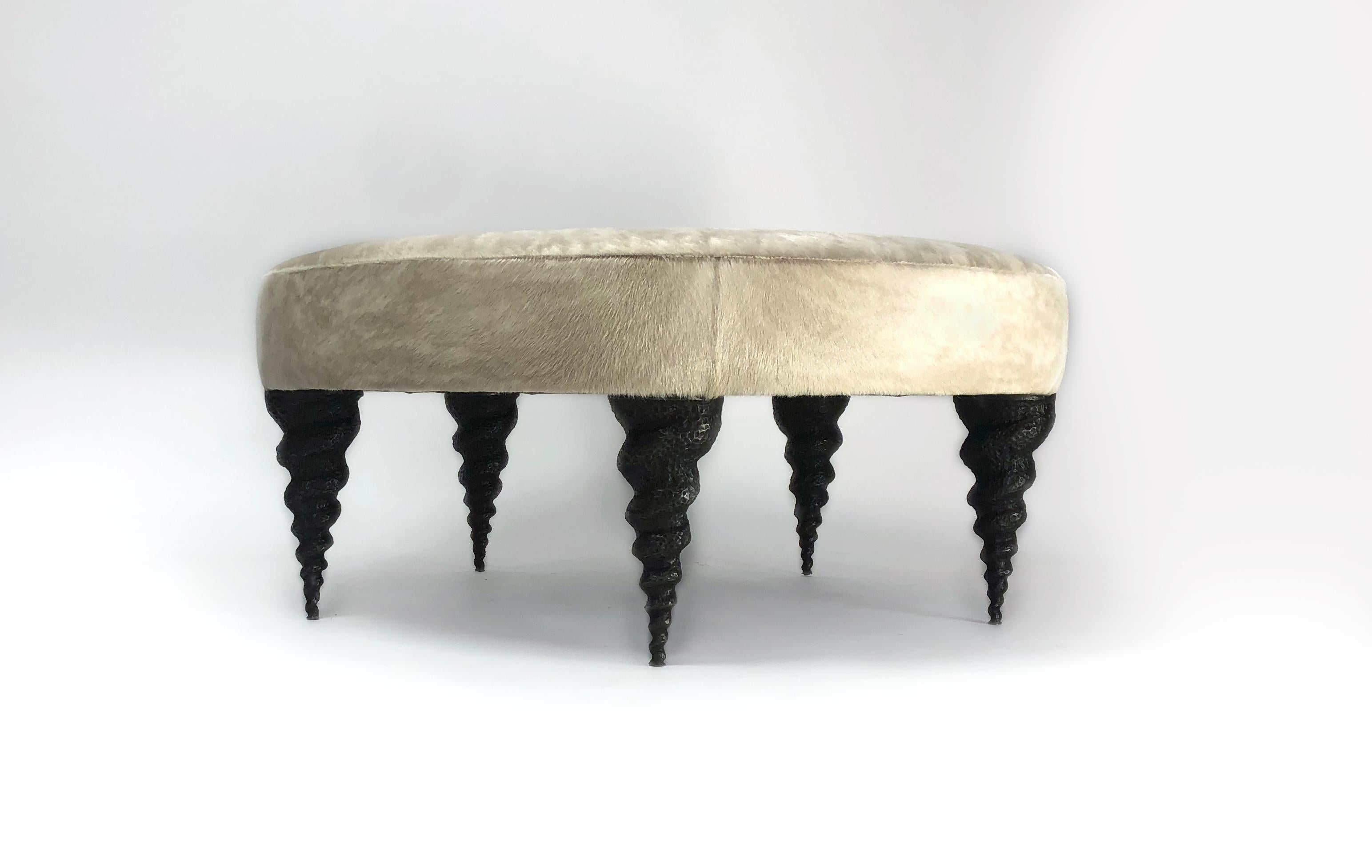 The legs of this ottoman have a biomorphic design. Cast in bronze, the five whimsical legs are inspired by Turrid sea shells. We upholster it to order with your fabric or hide.  Custom dimensions and shapes possible.  Perfect as a coffee table, or