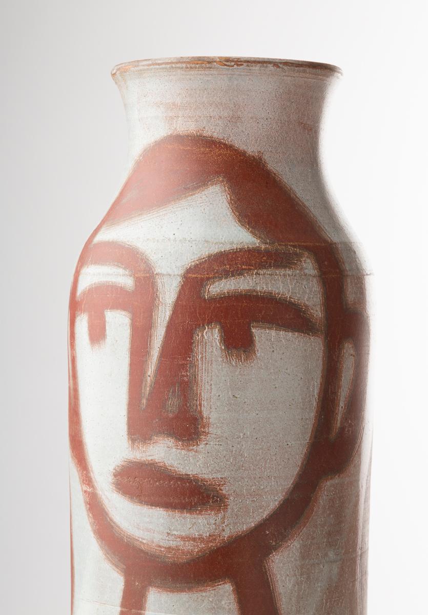 Este jarrón de cerámica de Robert Picault tiene forma cilíndrica con cuello estrecho y está decorado con una gran cara estilizada pintada en tonos rojos terrosos. Las líneas atrevidas y gráficas crean un llamativo contraste contra el fondo claro,