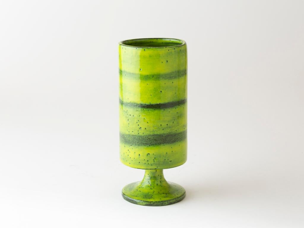 Este jarrón de gres de Mado Jolain, creado en la década de 1960, se alza sobre un pedestal en forma de cáliz y presenta un alto cuerpo cilíndrico. Su esmalte verde vibrante, animado por bandas horizontales más oscuras y un sutil moteado, refleja el