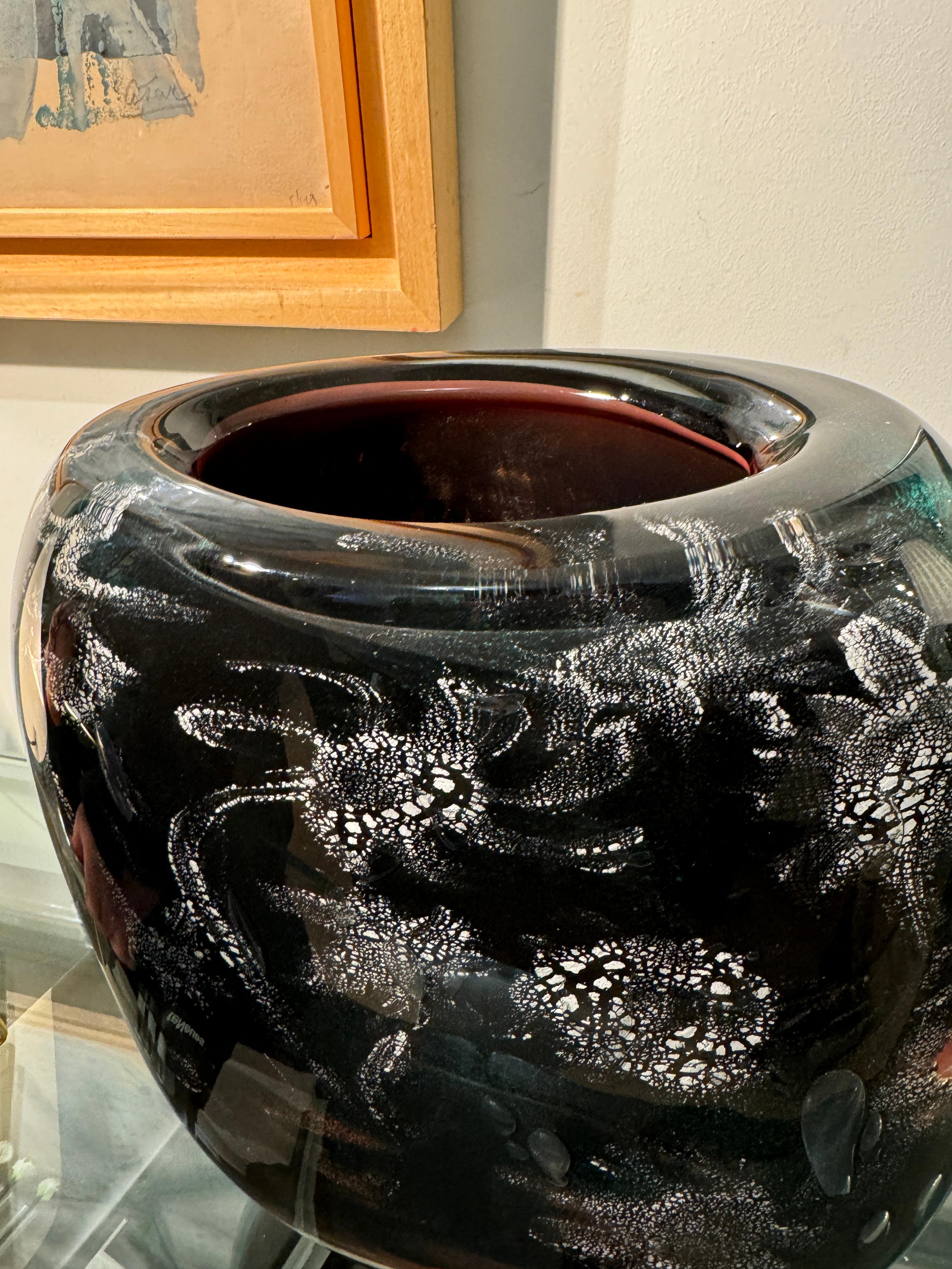 Superbe vase en verre soufflé signé Ada Loumani, artiste verrier renommé pour ses créations uniques avec des inclusions d’inspiration florale ou organique. Ce vase présente un magnifique jeu de textures et de couleurs sombres avec des motifs blancs