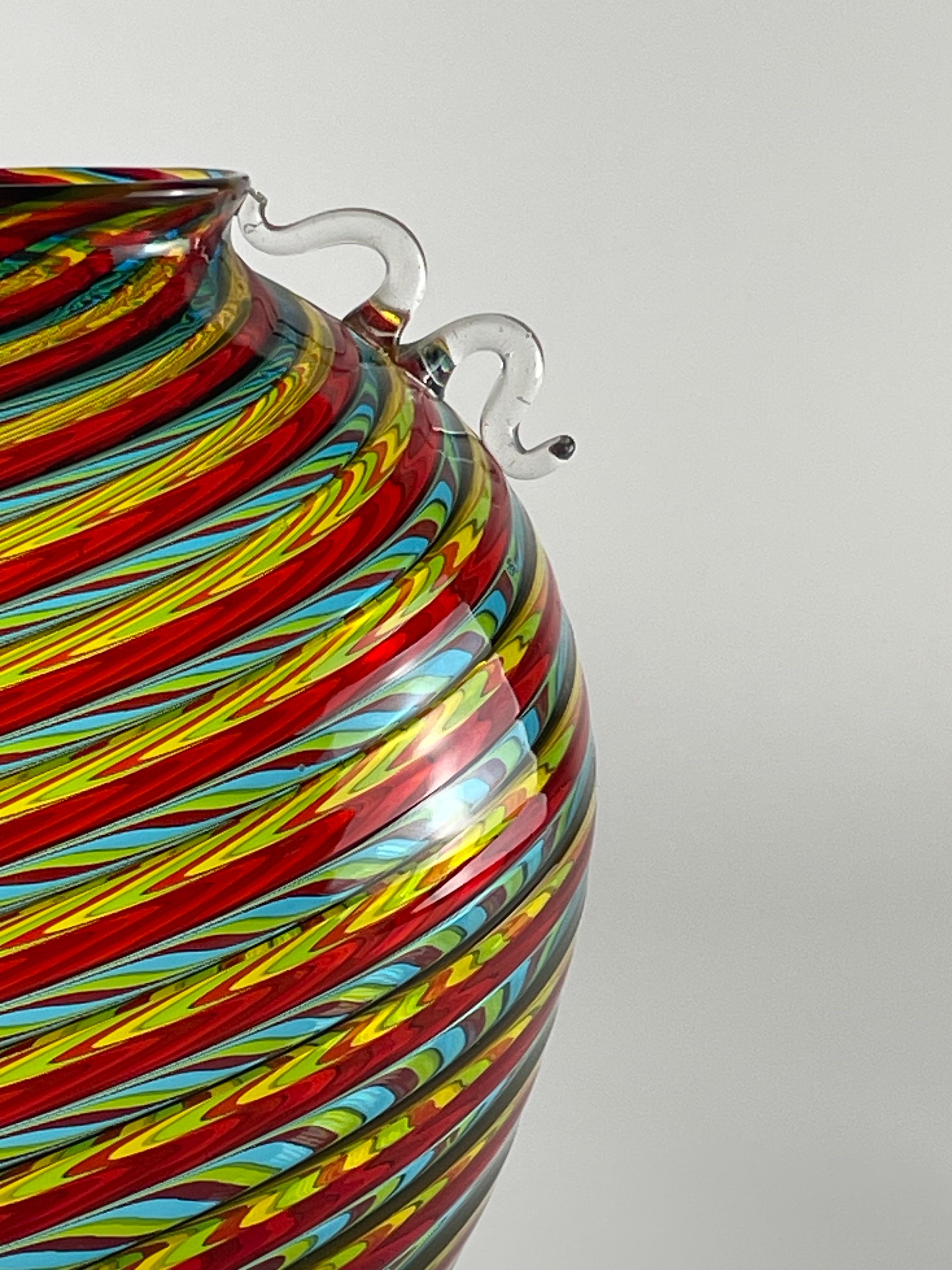 Grand vase coloré composé de tiges de verre rouges, jaunes et bleues alternées. Les poignées supérieures, sur les deux côtés, sont en verre de cristal, travaillé et appliqué à chaud sur le corps du vase. Fabriqué à Murano par le maître Bruno