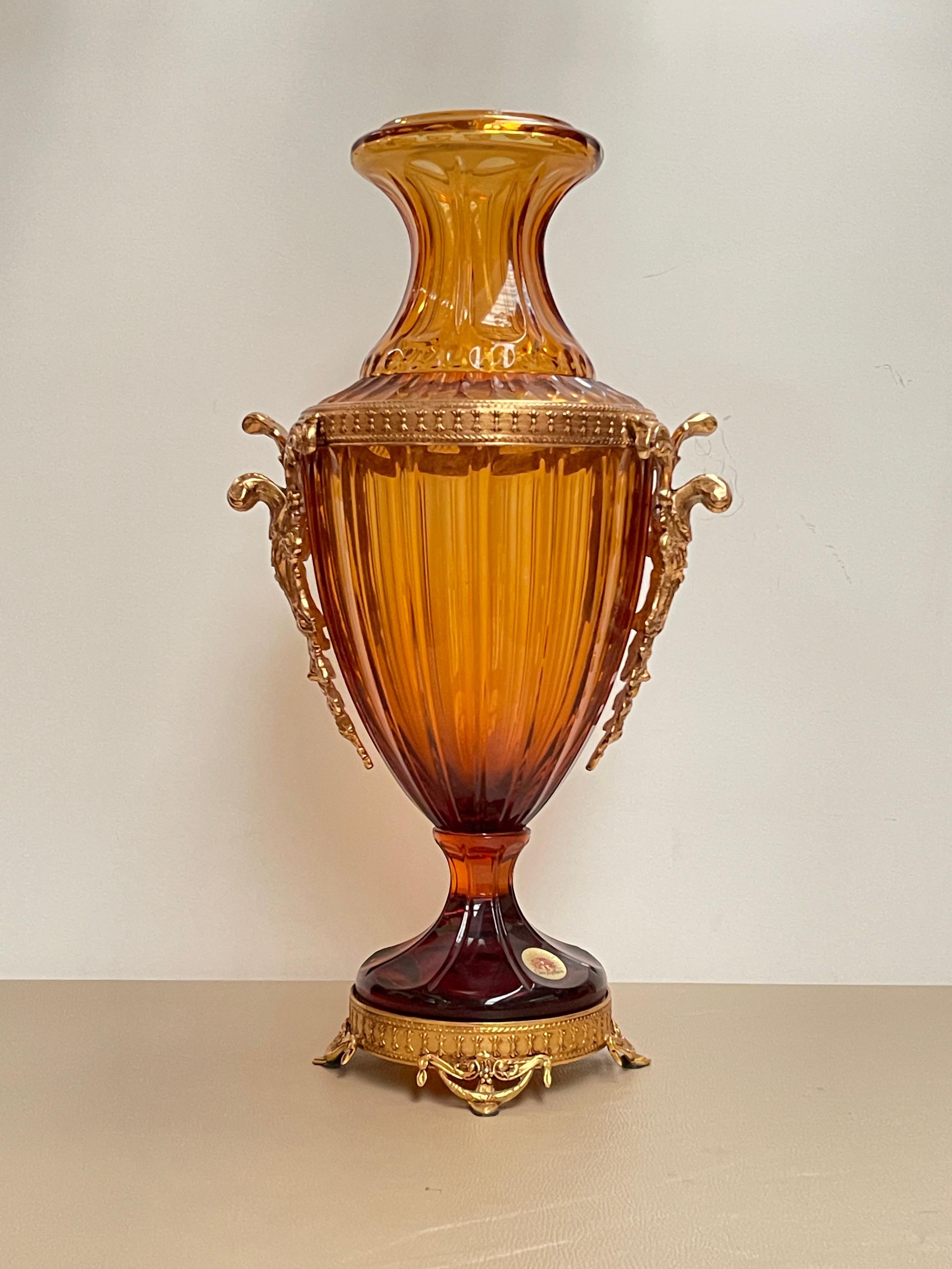 Raffinato vaso in cristallo soffiato di colore ambra sfumato, impreziosito da una montatura in bronzo dorato cesellato. Il design elegante richiama lo stile neoclassico francese, con una forma slanciata, orlo svasato e manici laterali finemente
