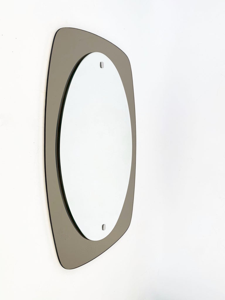 Wonderful midcentury oval wall mirror with bronzed glass frame. This elegant and charming two levels piece was produced by Veca during the 1970s in Italy.

It features an oval grey mirror and a bronzed nickel-plated frame with brass screw heads