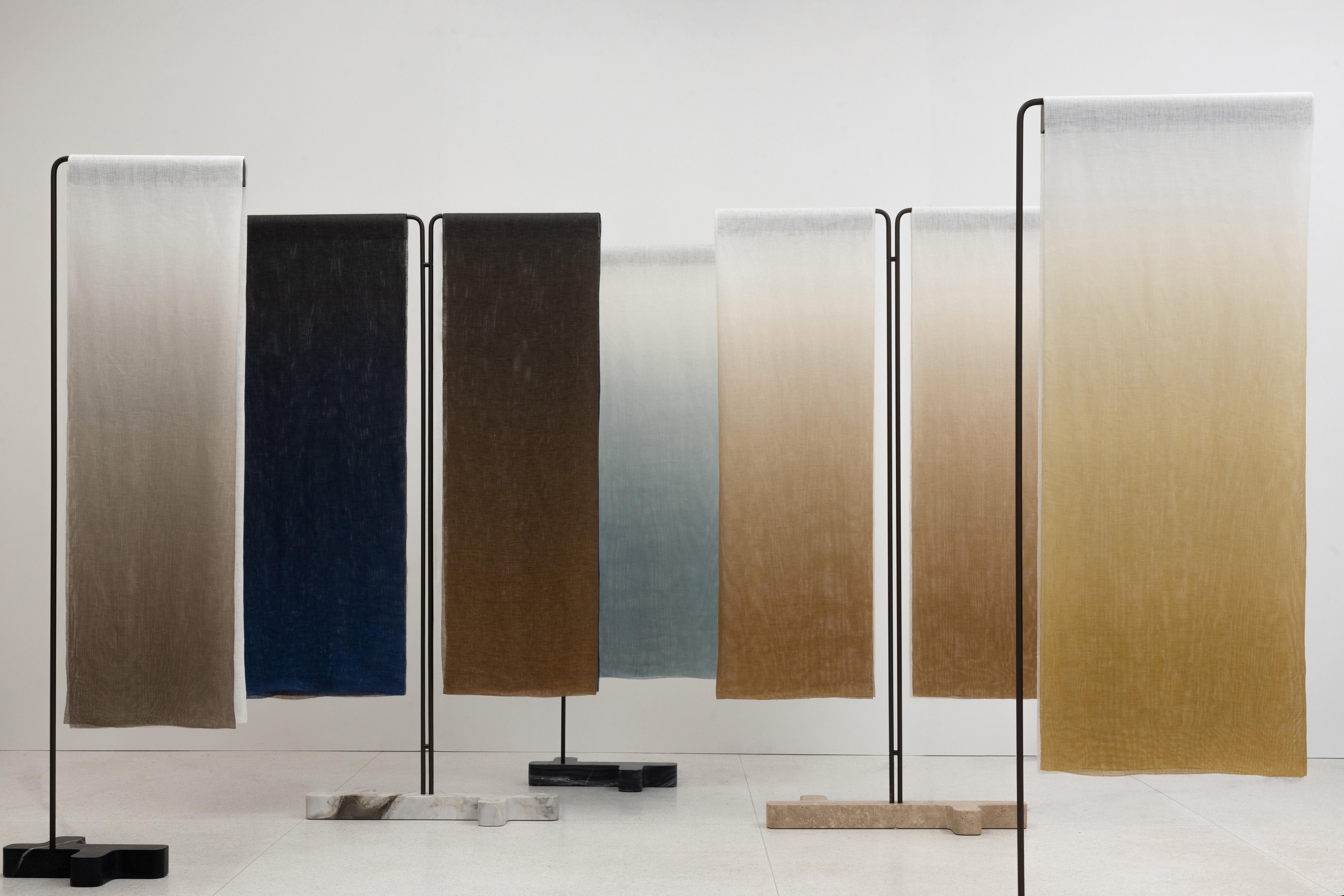 The fabric in this design transcends its traditional role as a simple covering and becomes a narrative element, engaging both light and movement. Resting on a slender metal frame supported by a marble base, rectangular pieces of gradient-dyed linen