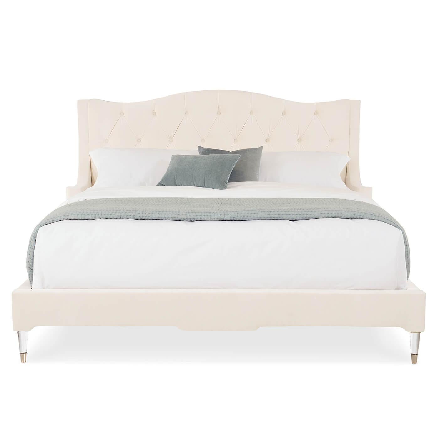 A velvet tufted upholstered king bed with a tufted diamond-pattern headboard. This bed is upholstered in luxurious cream velvet. The sides of the headboard wrap around with a design inspired by an English armchair. It has wooden legs finished in