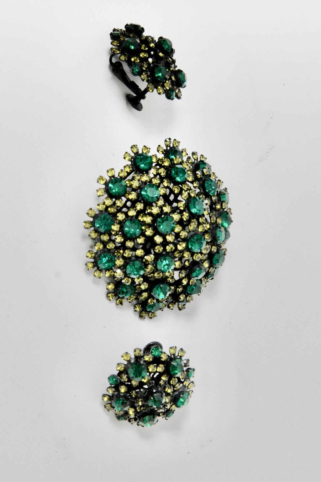 Vendome semi-parure of dome-shaped brooch and clip-on earrings composed of large round green crystals, each surrounded by small lime-colored crystals in a floral manner, and each with a filigree frame of black metal. The earrings have adjustable