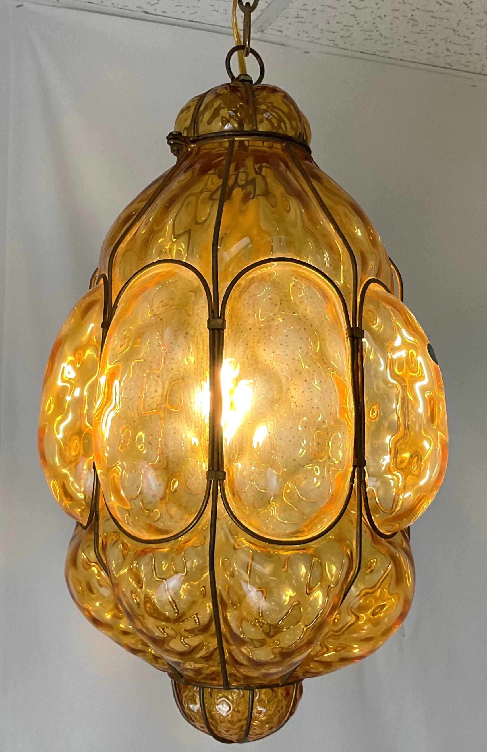 A striking 1970s Italian Murano glass pendant featuring a richly textured amber glass shade enclosed in a sculptural brass frame. The organic, handblown form displays a mesmerizing pattern of molten swirls and undulating contours, creating a