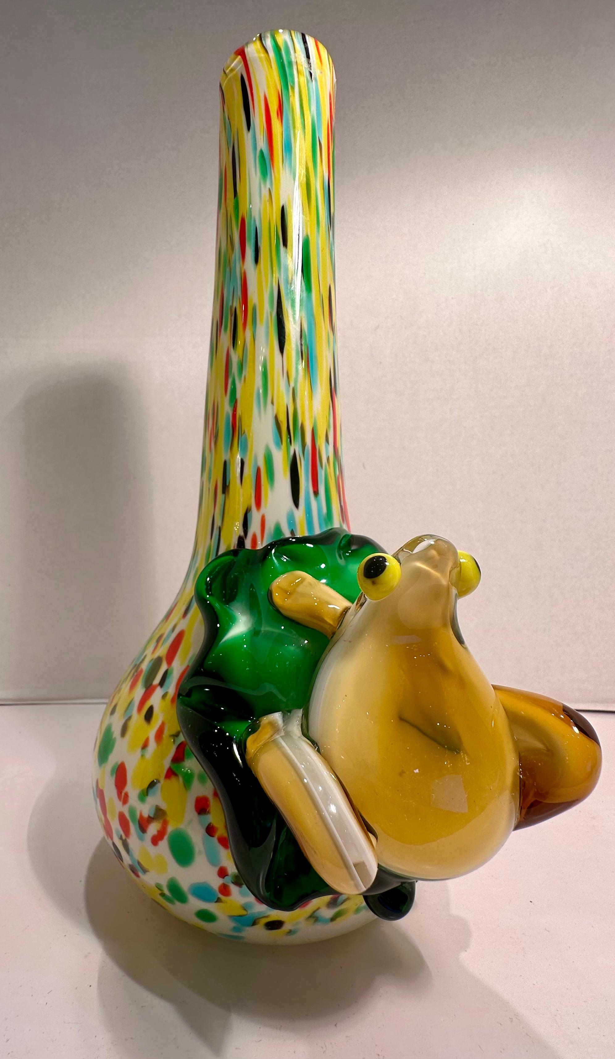 Molto artistico, stravagante e colorato, realizzato a mano e soffiato a bocca in Italia, il vaso a collo di bottiglia multicolore con fondo a bulbo presenta una rana in vetro artistico giallo oro, dal ventre bianco, su una ninfea verde affiancata