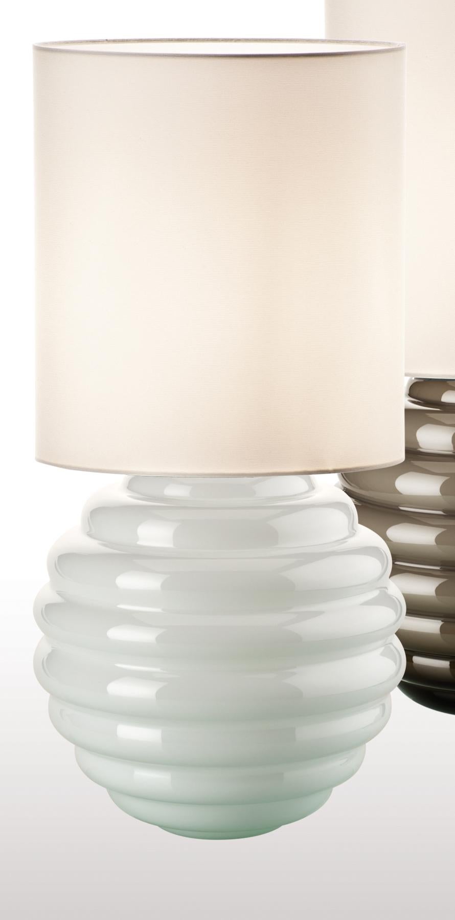 Deco table lamp in glass with a white fabric shade. Its body's rippled shape make it an interesting addition to any living room, office or bedroom space. Also available in other colors.

Light source: One max 105 E27. Dimensions: 30 cm diameter x