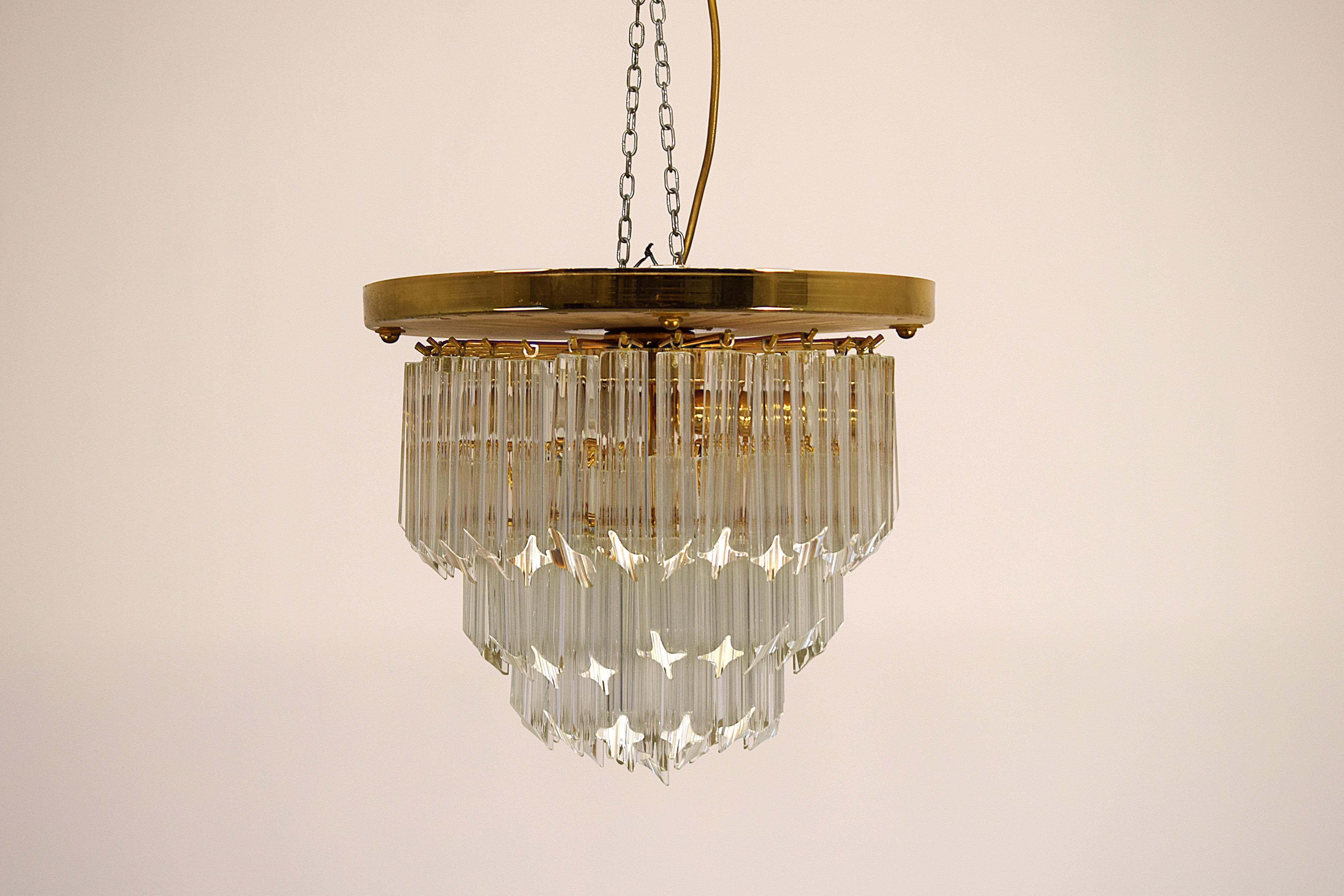 Elegante lampadario Venini originale degli anni '70, realizzato in vetro e metallo dorato. Questo raffinato pezzo di design vintage italiano è composto da elementi in vetro sfaccettato disposti su più livelli, che creano giochi di luce suggestivi.