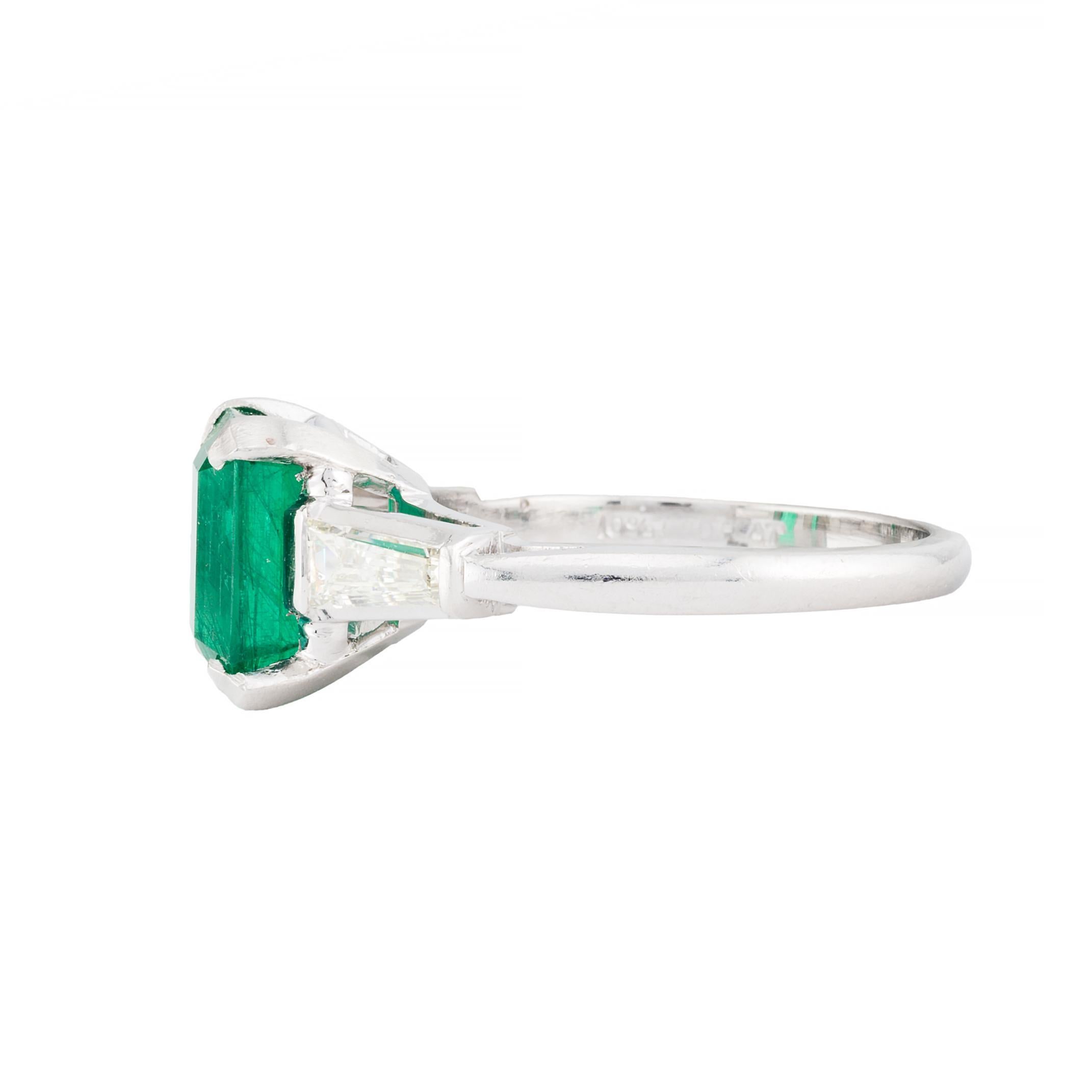 Aston Estate Jewelry Presents:

A striking emerald and diamond three stone ring featuring a 2.52 carat emerald cut emerald of vibrant green color. The emerald is elegantly set in a minimalist design, accentuating its natural beauty. Flanking the