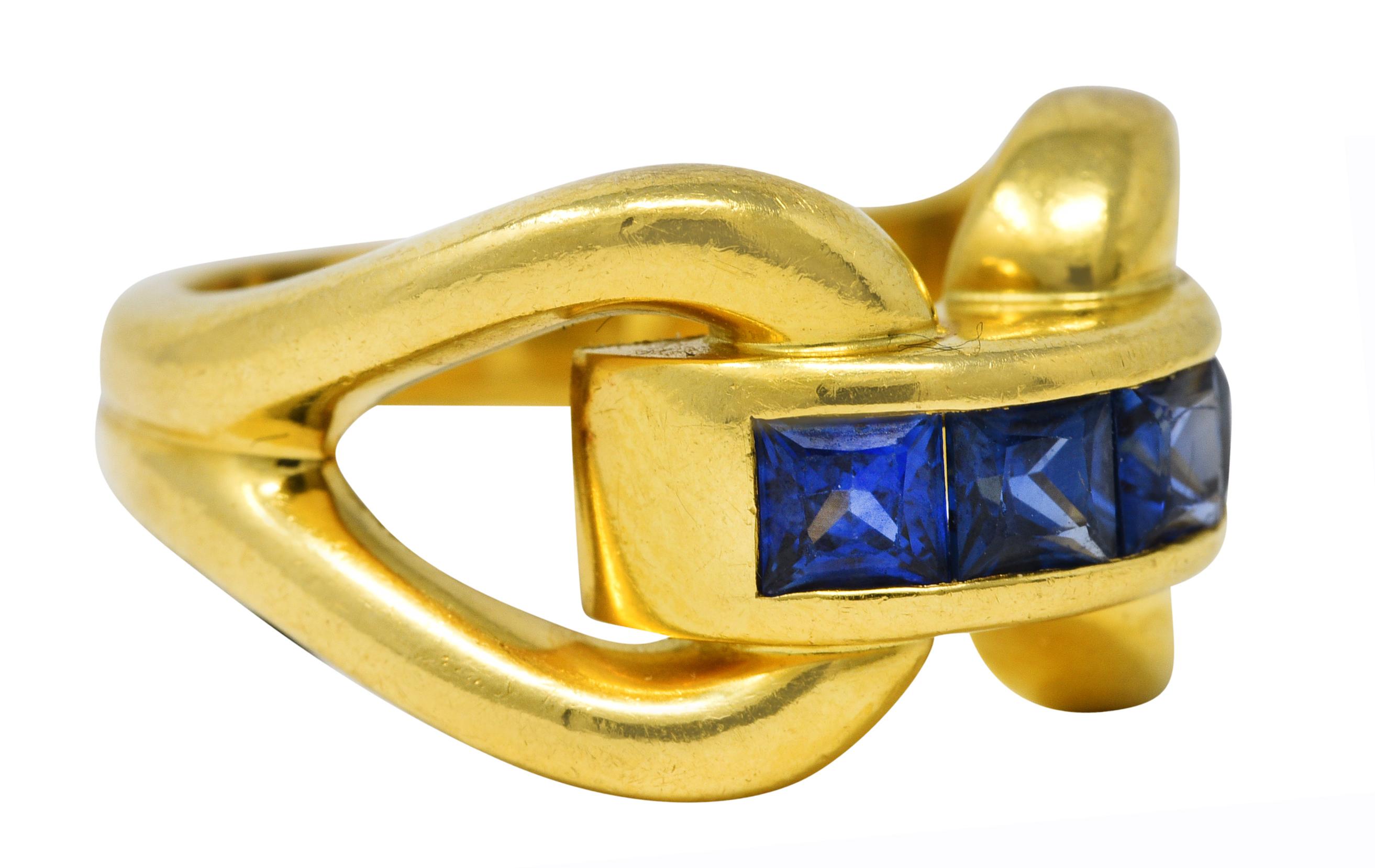 Ring features four square cut sapphires channel set to front - weighing approximately 0.88 carat total. Transparent and deeply saturated blue in color. Flanked by stylized horsebit motif gold shoulders. Completed by grooved shank. Stamped 750 for 18