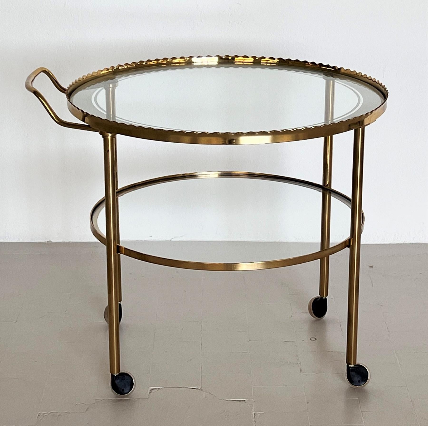 Beautiful elegant serving trolley or bar cart from the 1960s with four wheels.
The beauty of this trolley is the form of the upper brass frame.
The two trays are made of glass; the upper glass plate has a clear creamy white stripe on the outer