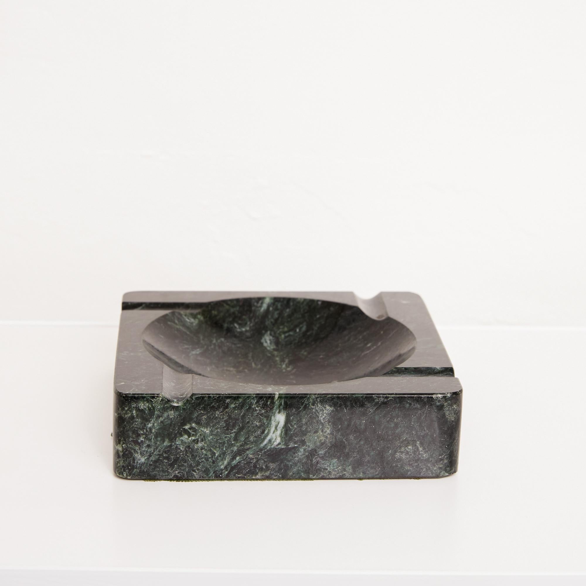 A square ashtray in a polished black marble with green and white veining by the Vermont Marble Company of Proctor, VT. The piece has a circular well, and four routed asymmetrical indentations perpendicular to its edges. The corners and edges of the