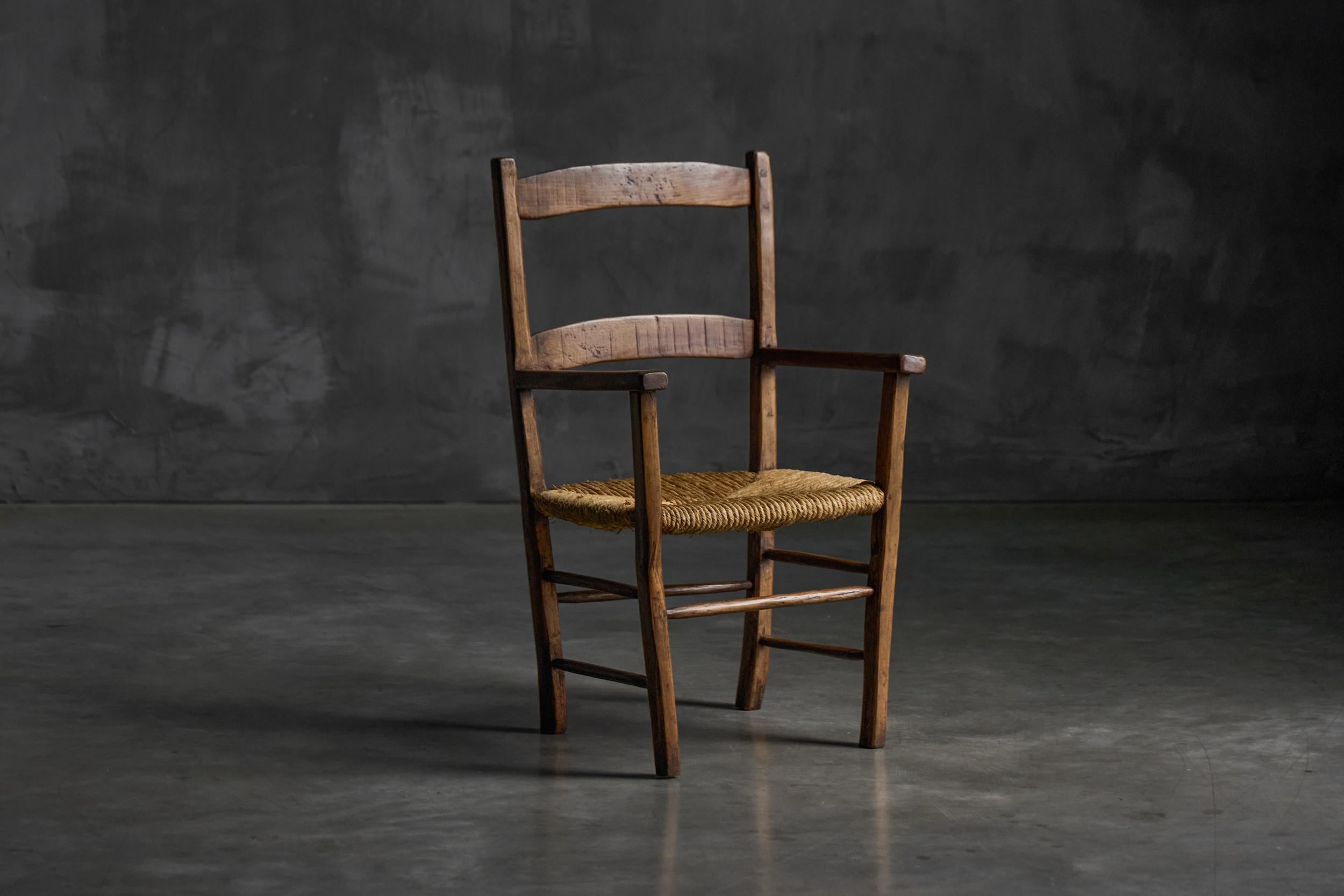 Este sillón alsaciano del siglo XIX presenta un armazón de madera teñida de oscuro, típico de la artesanía popular regional. El respaldo incluye dos listones horizontales rematados con remates redondeados, que le añaden un encanto rústico. Los