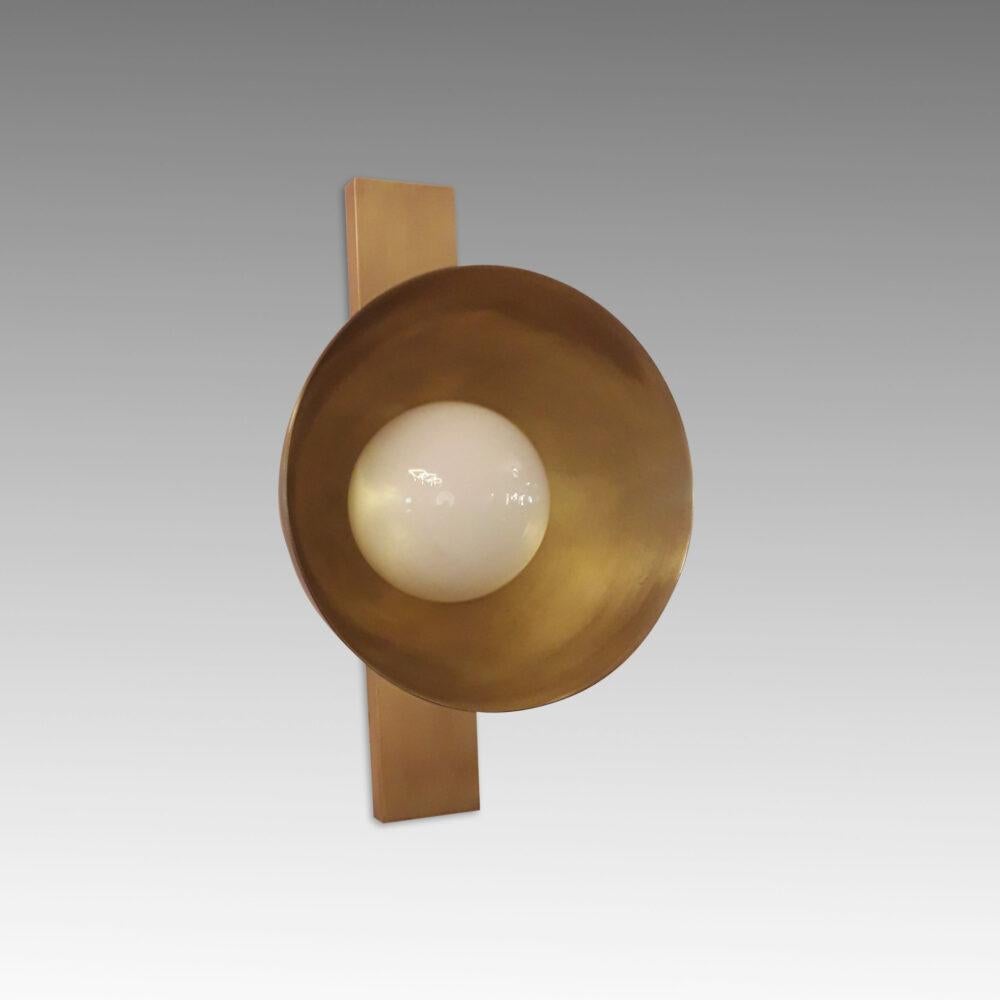 L'applique Verner 1 combina due semplici forme geometriche: un paralume emisferico rivolto in avanti e una piastra posteriore rettangolare verticale, entrambi realizzati in ottone patinato oro invecchiato. Il contrasto tra la curva della cupola e le