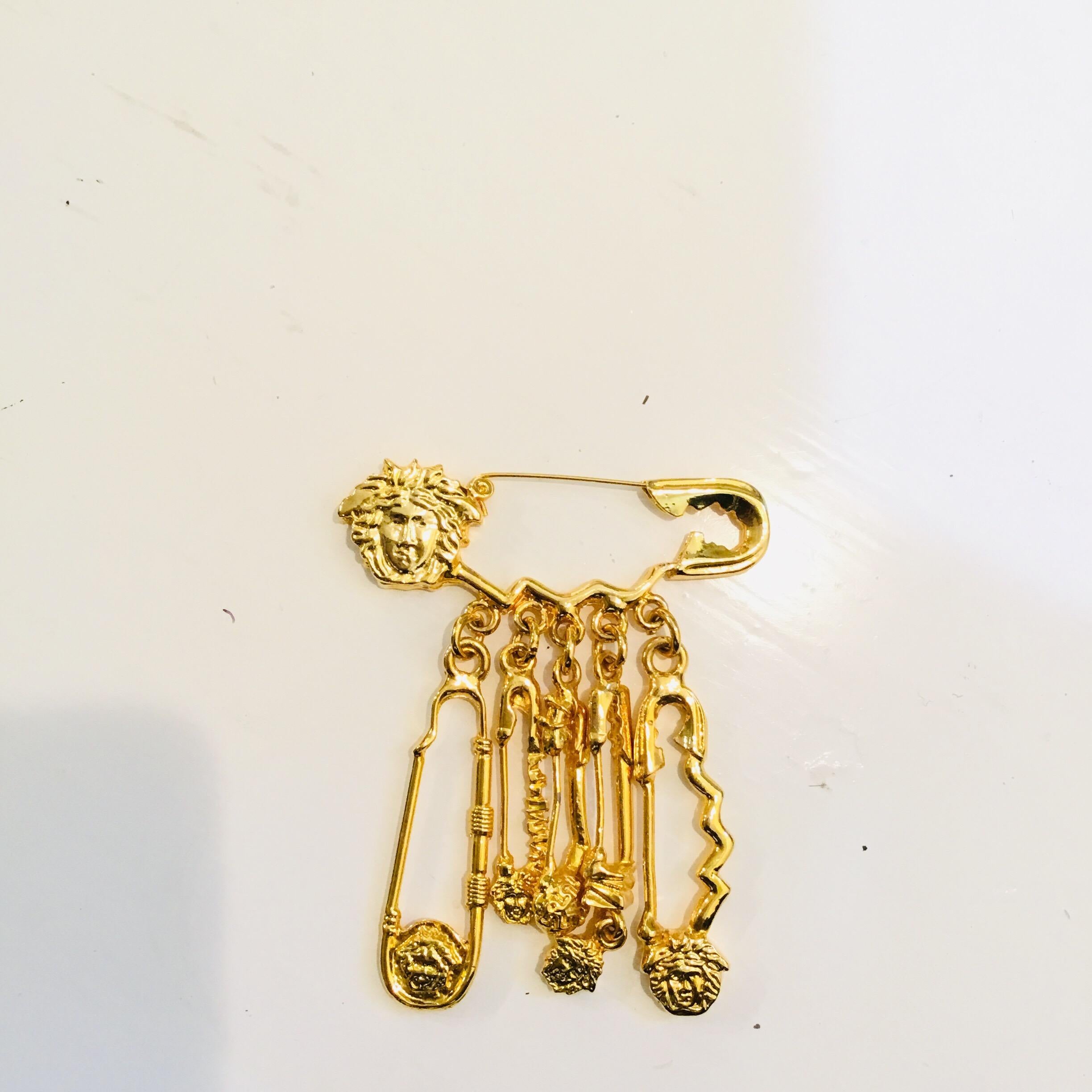 Versace gold brooch features a signature safety pin design with 5 dangling pins with Medusa head detailing. Brooch measures 2.5 inches across and 3.25 inches long. Brooch is unmarked, 100% authentic.