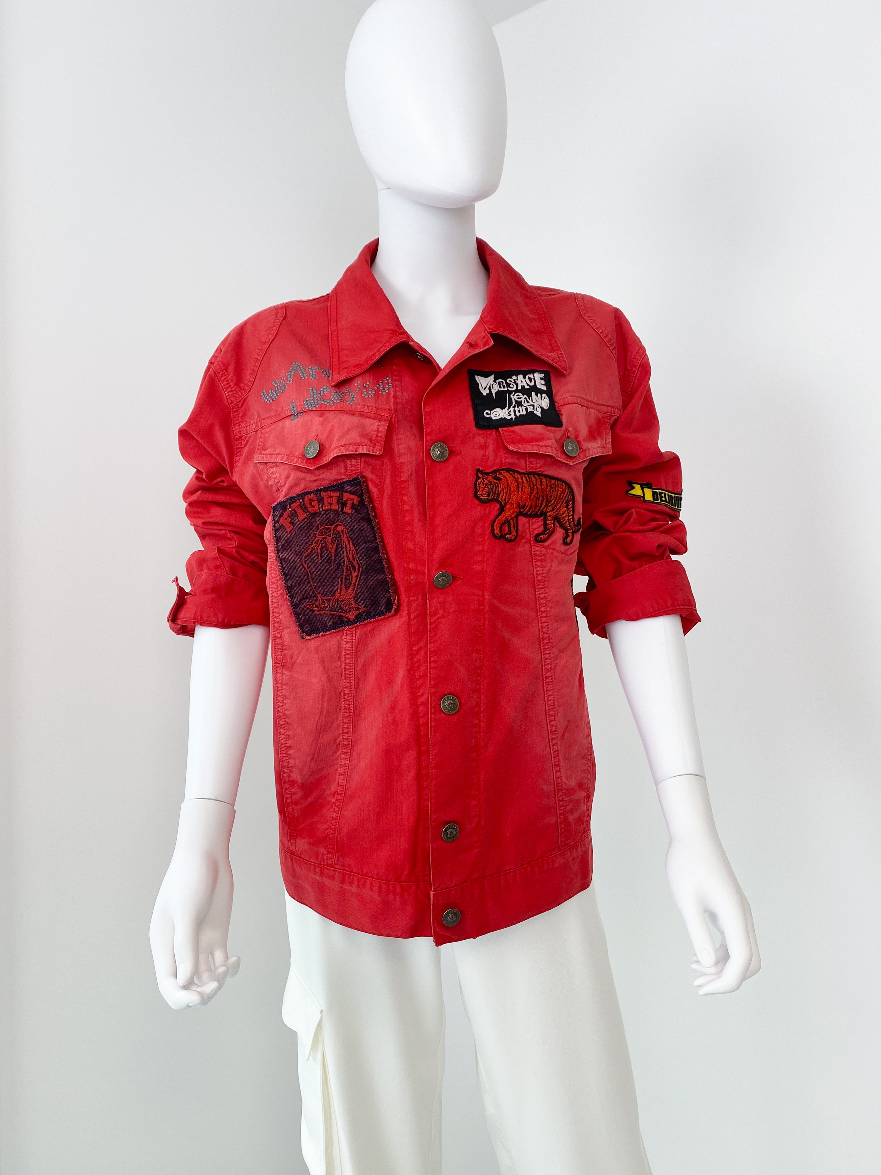 A rare red denim jacket by Versace Jeans Couture from the Delirium Collection. Unisex style, it will fit men size XL and women size XL. Embroidery, graffiti, and patches are all over the jacket. The jacket has long sleeves with button cuffs and