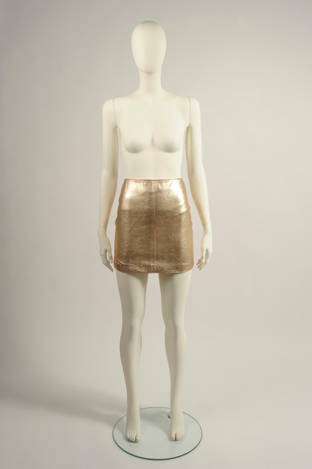 Dieser Minirock ist Teil des Looks Nr. 58 der Herbst-Winter-Kollektion 1994 von Versace. Er ist aus goldfarbenem Leder mit Eidechsenoptik gefertigt (siehe Bilder 7 & 8). Die italienische Größe 38 (US 0) ist vollständig mit Viskose gefüttert und
