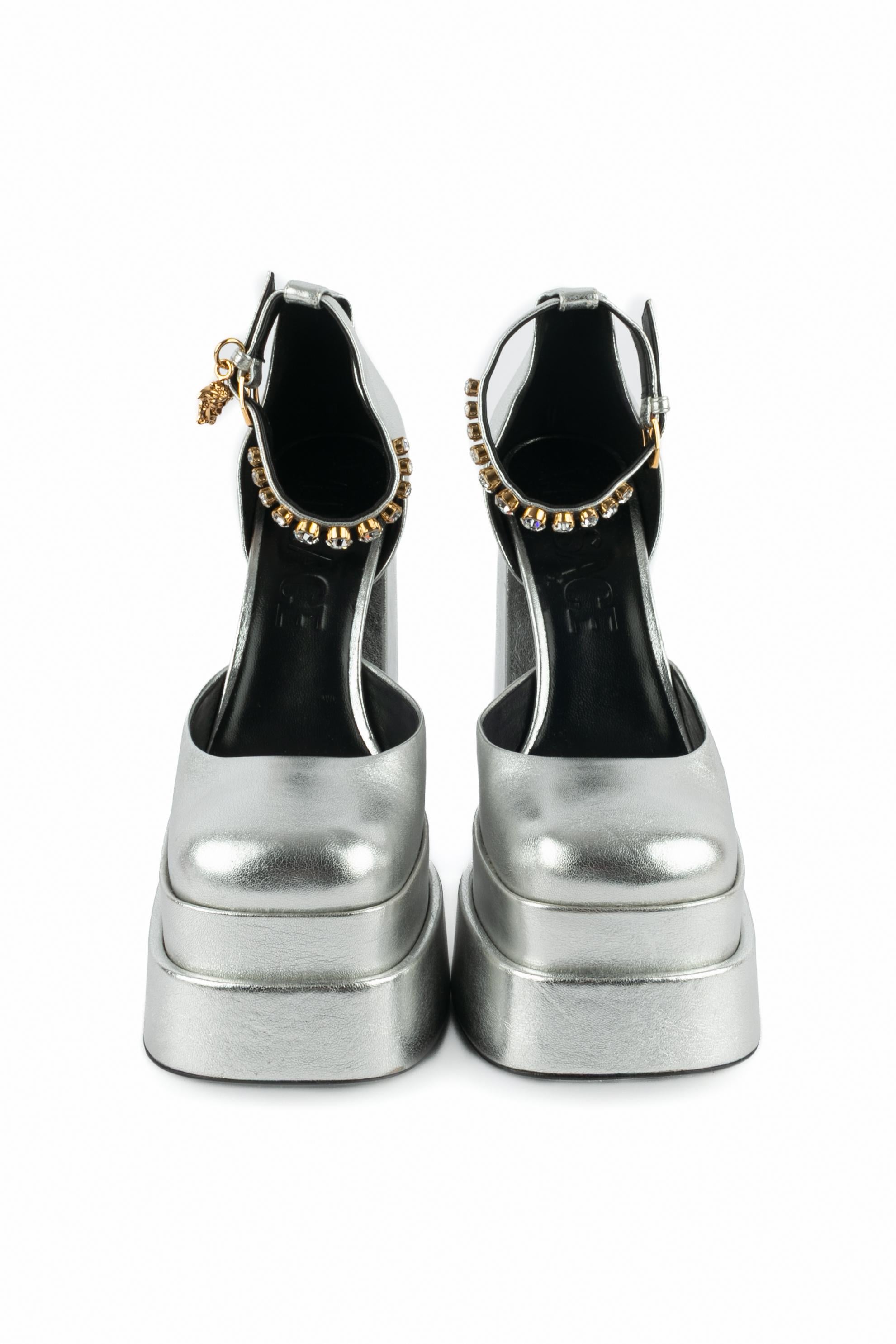 VERSACE - (Made in Italy) Hochhackige Stöckelschuhe aus silbernem Leder mit zweistufiger Plateausohle. Die Handgelenkszaumzeugs sind mit rosa Strasssteinen verziert und mit goldenen Metallcharms in Form von Medusen verziert. Größe 40FR angegeben.