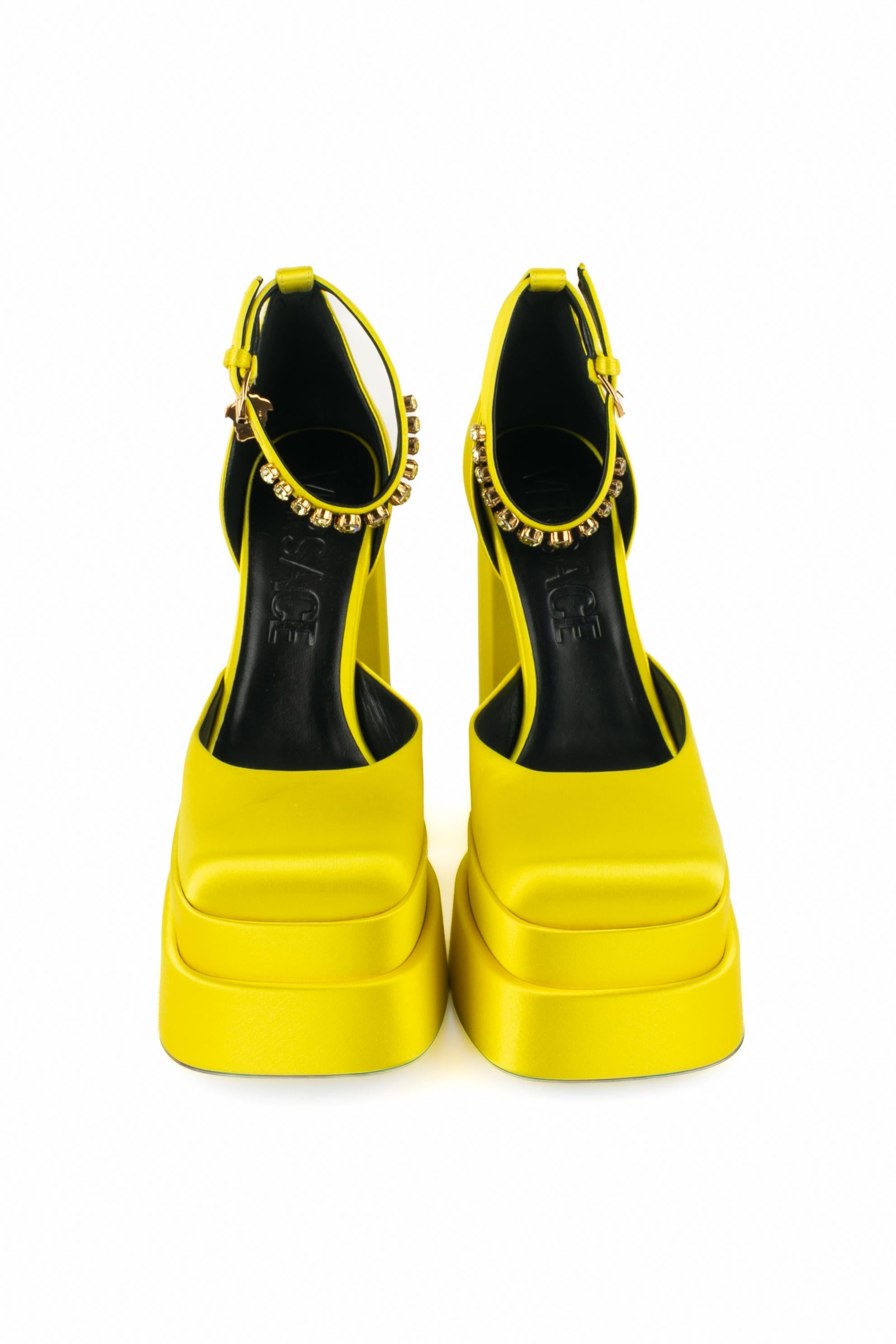 VERSACE - (Made in Italy) Gelbe Stöckelschuhe mit Blockabsatz aus Satin und zweistufiger Plateausohle. Die Handgelenkszaumzeugs sind mit gelben Strasssteinen verziert und mit goldenen Metallcharms in Form von Medusen verziert. Größe 40FR angegeben.