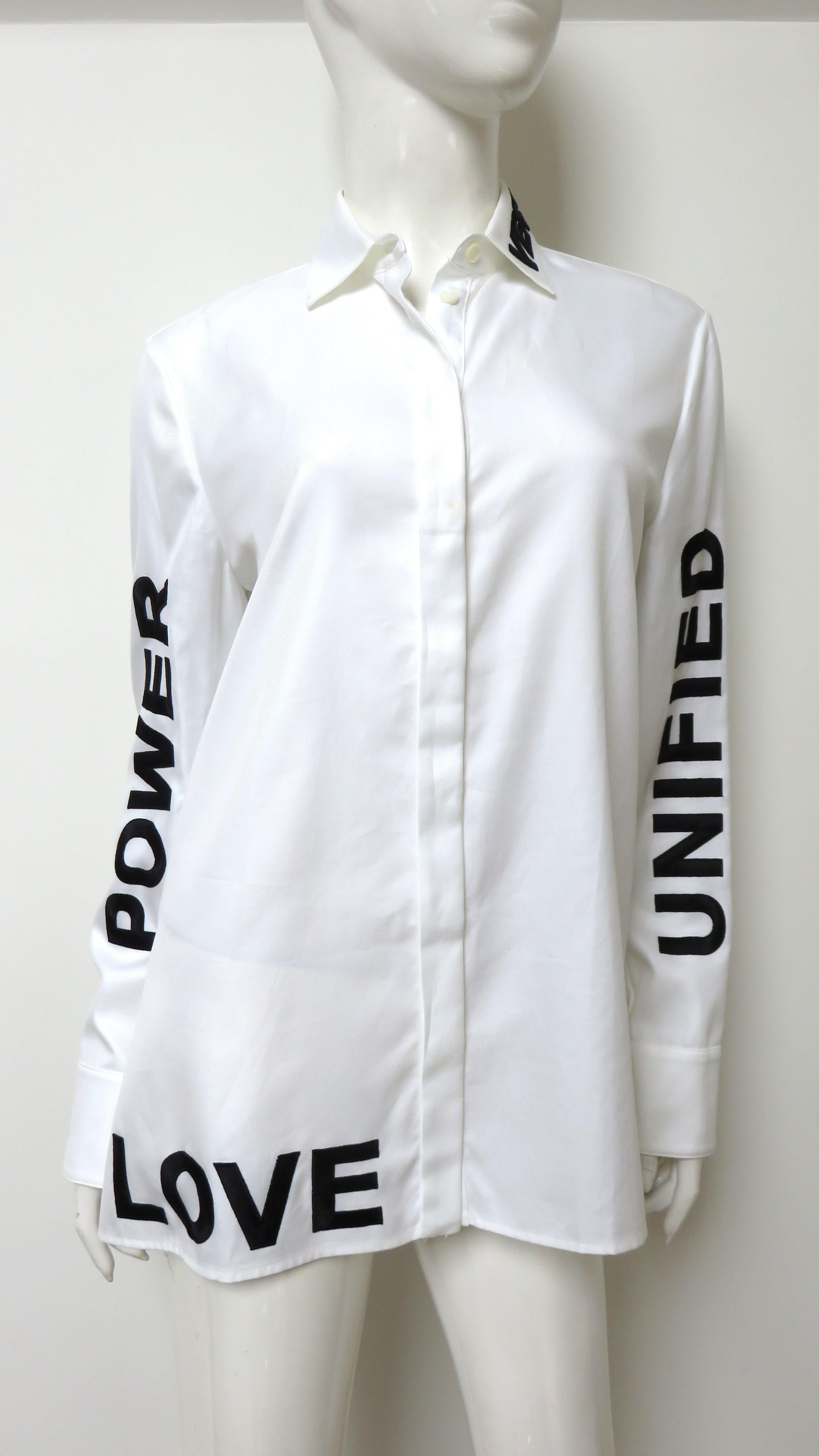 Three words on this white shirt from Versace- Love, Unified and Power.  The words are appliqued in black silk capital letters along each sleeve and the bottom front left hem.  Letters spelling Versace are along one side of the collar. It has Versace
