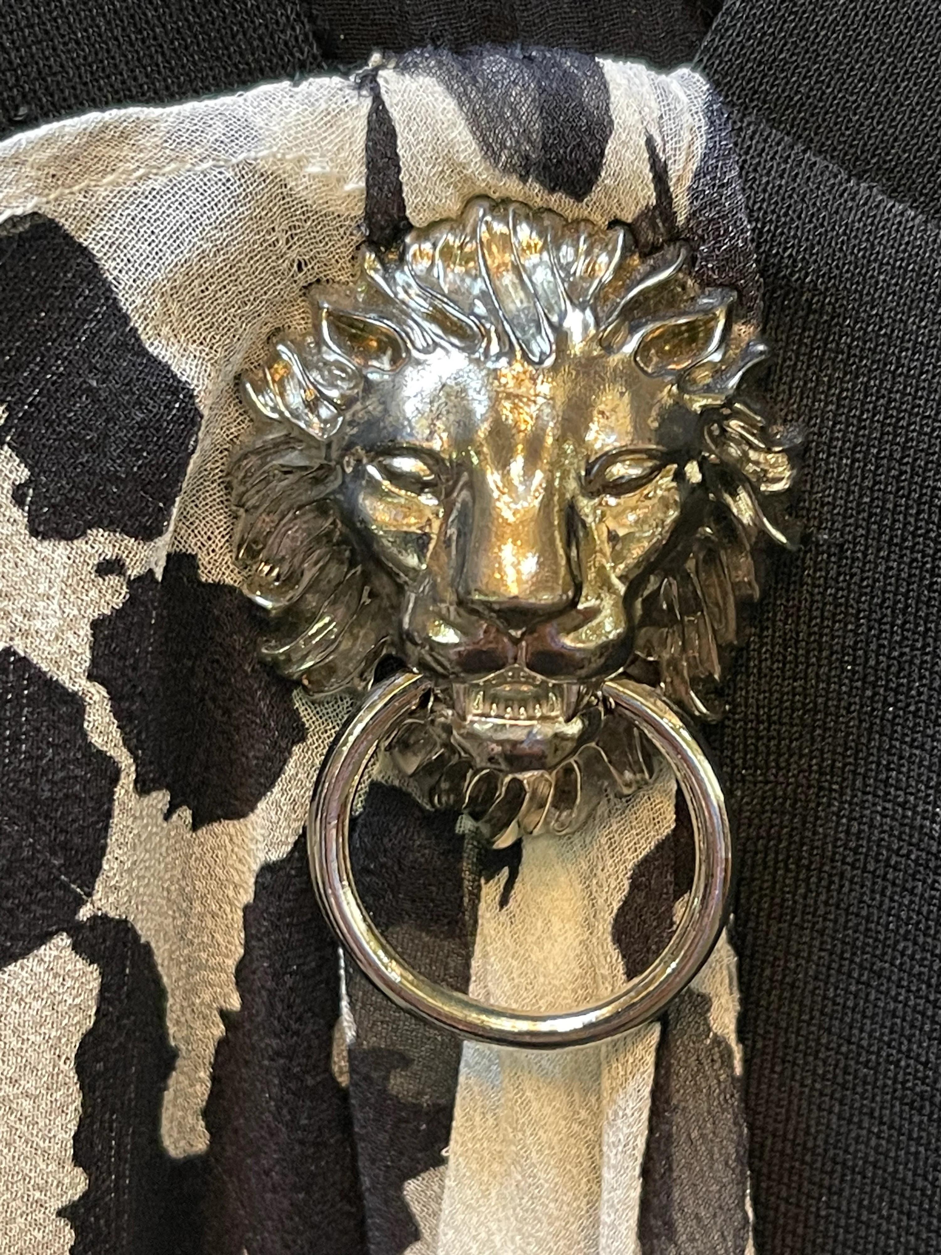 2016  mayhem from the House of Versace. Black extreme body con mini dress with side cut outs. Deep (to your belly button) v neckline. Lions head logo in chrome at waist.  Giant swath of white chiffon emblazoned with stylized logo floats and shimmies