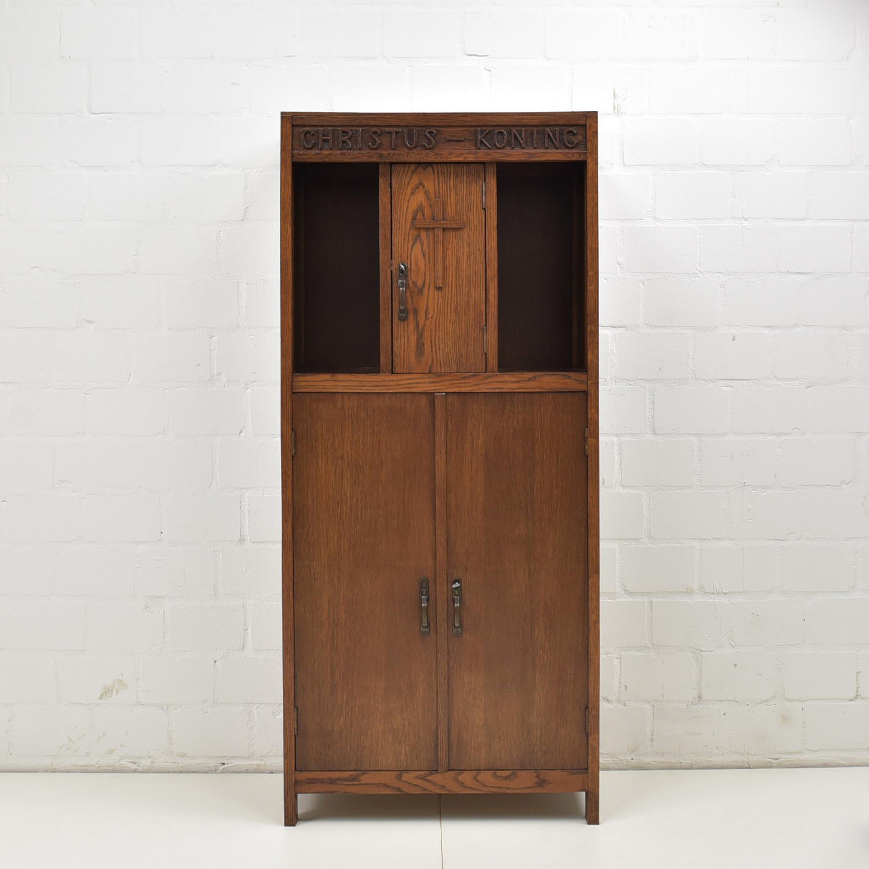 Narrow Christian cabinet restored 1930 Oak Vertiko Christkönig

Features:
Narrow model with two doors and three shelves below and three compartments, one with a door, above
Upper door with Jesus cross
Carved inscription 