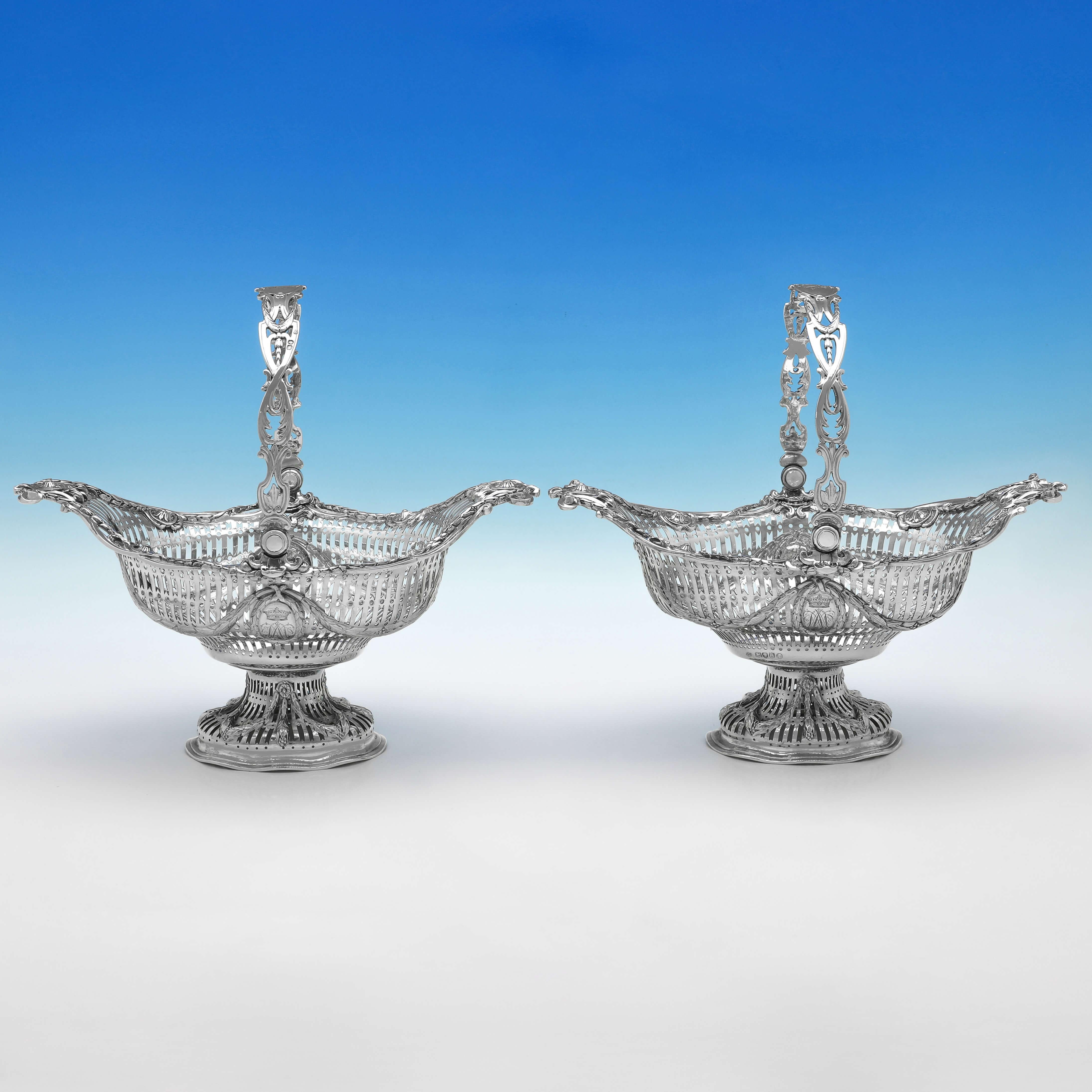 Hallmarked in London in 1882 by George Fox, this striking pair of Victorian, Antique Sterling Silver Baskets, are ornately decorated, with pierced sides and feet, cast and applied rosettes and swags, pierced swing handles, and engraved crests and