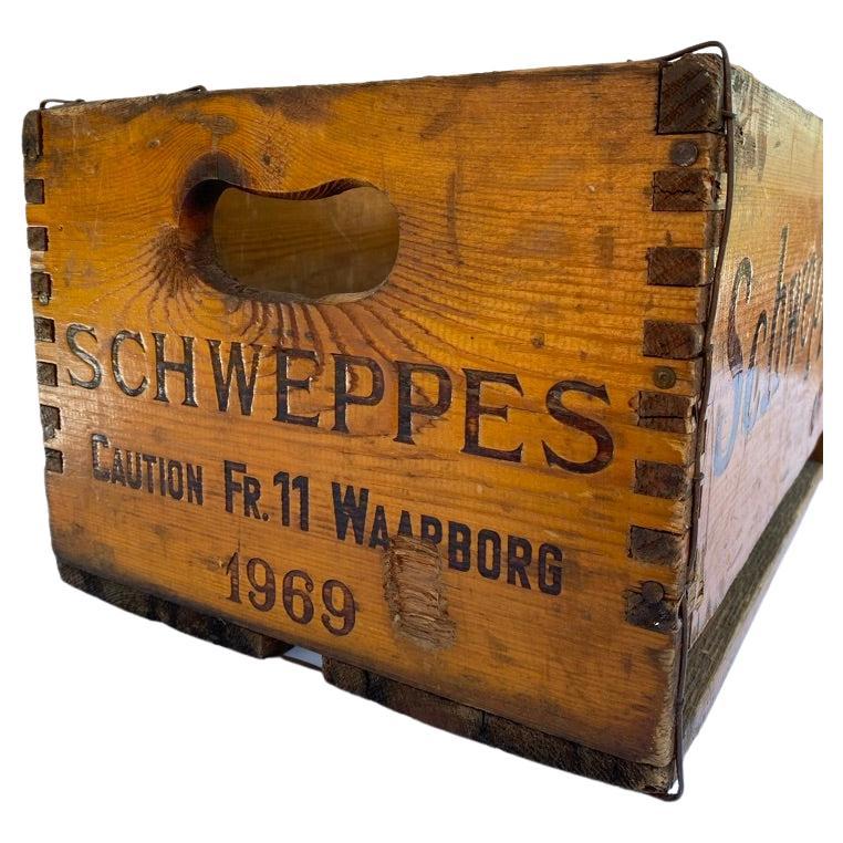 A 1960’s wooden Schweppes crate. Reads “SCHWEPPES Caution FR. 11 Waarborg 1969” on the short sides in black lettering, which means 11 Belgium Francs deposit, with oval cut out handles. “Schweppes ” on the long sides. Metal straps support the corners